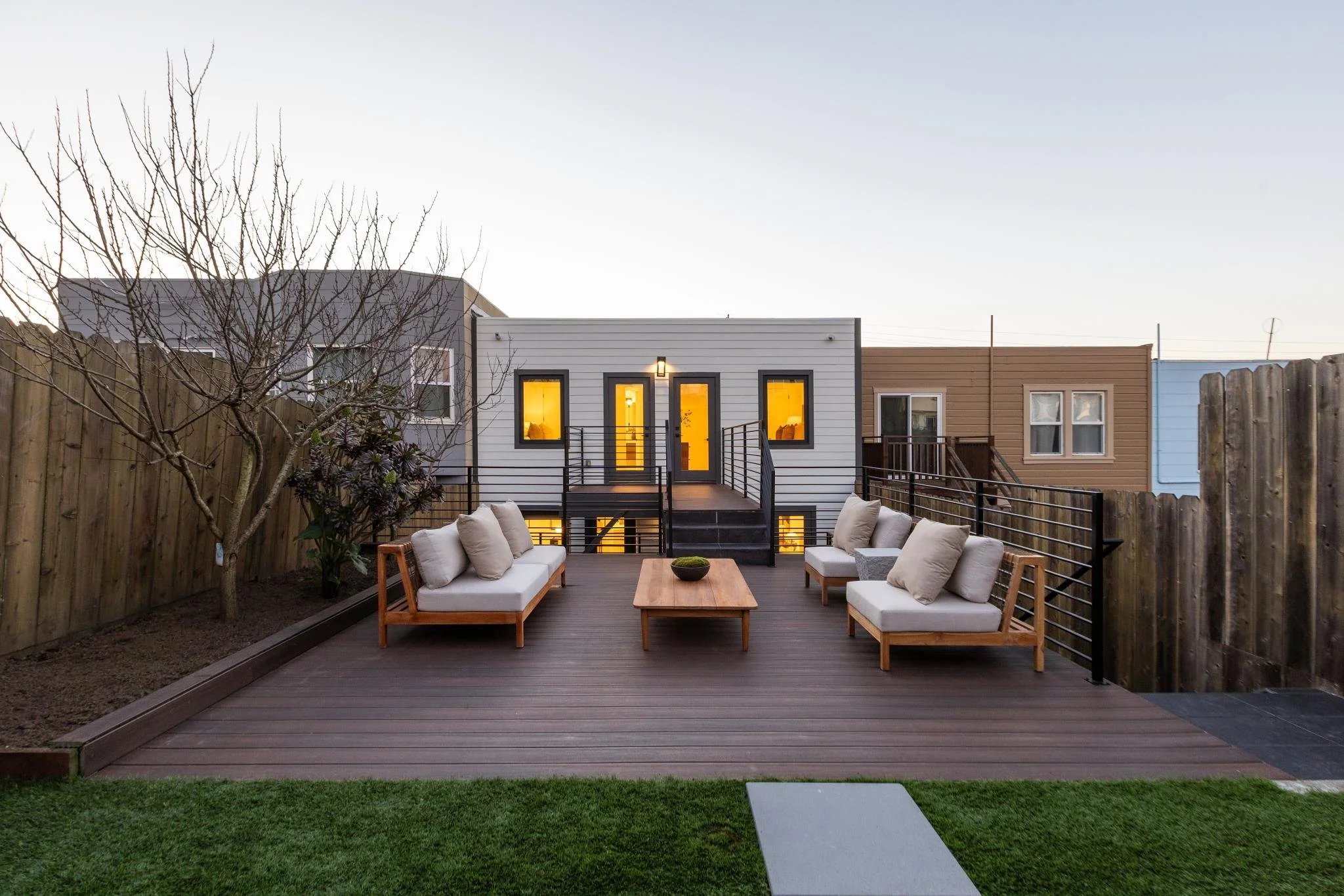 Backyard patio seating area with wooden deck, outdoor sofas with cushions, a wooden coffee table, fenced yard, and small trees during dusk.
