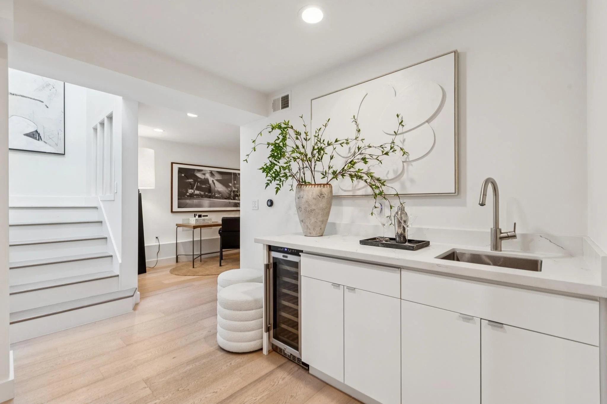 Contemporary interior with white cabinetry, a bar refrigerator, a large vase with greenery, artwork, and a sitting area in a well-lit living space.