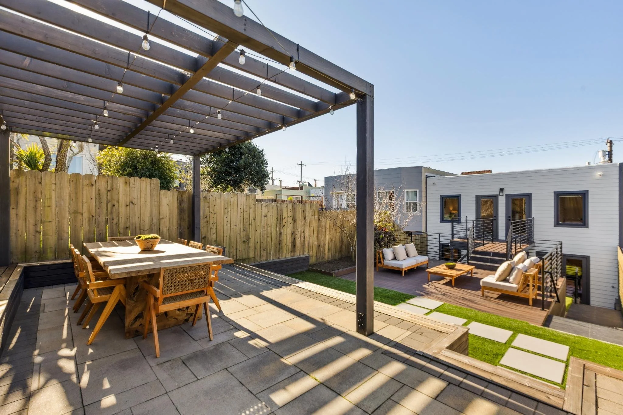 A backyard patio with a wooden pergola, outdoor dining table, and seating area with couches and chairs, surrounded by a wooden fence and neighboring house.