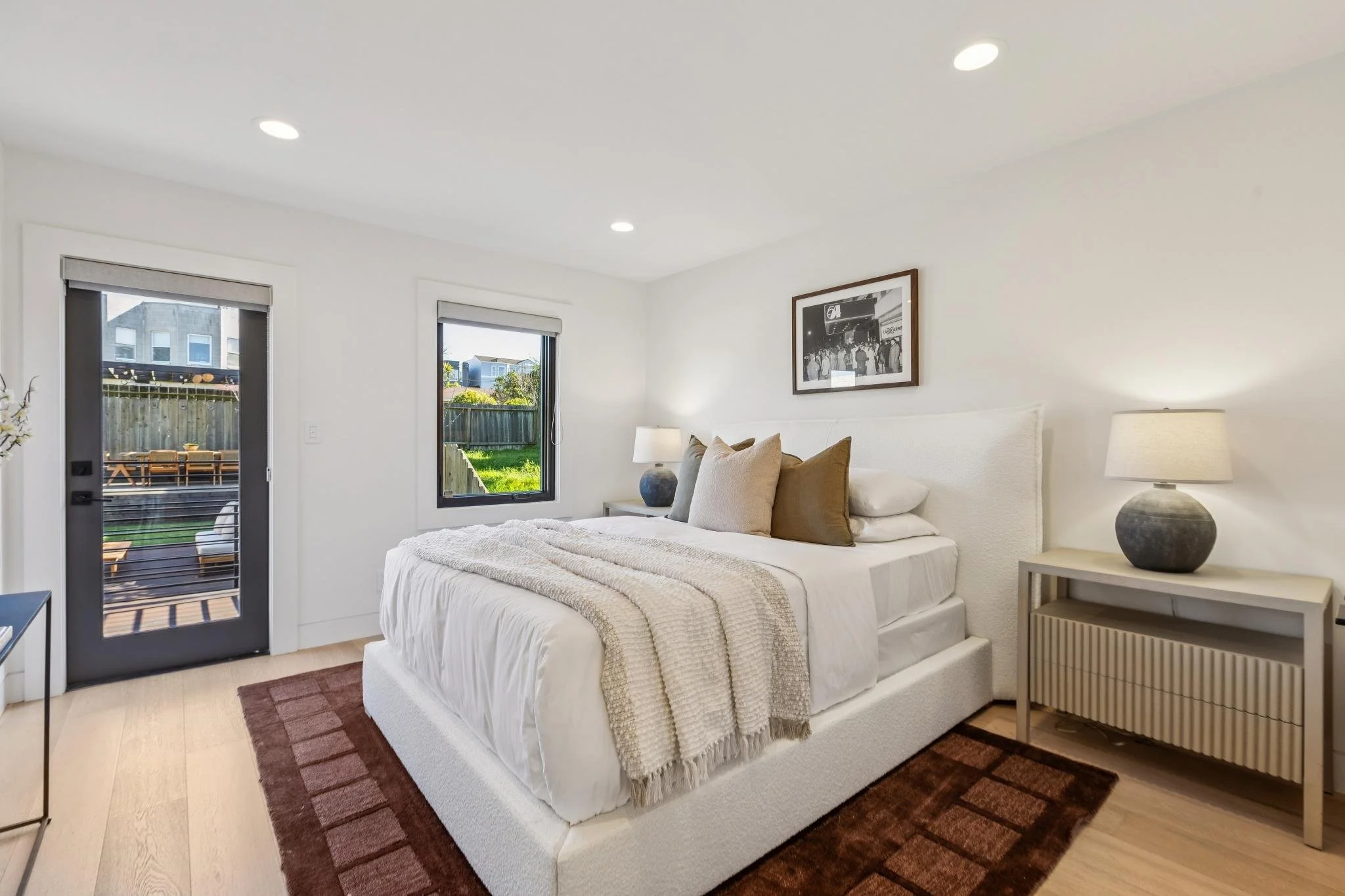 Bright bedroom with white walls, hardwood flooring, a white bed with decorative pillows, a nightstand with a lamp, and windows showing outdoor greenery.