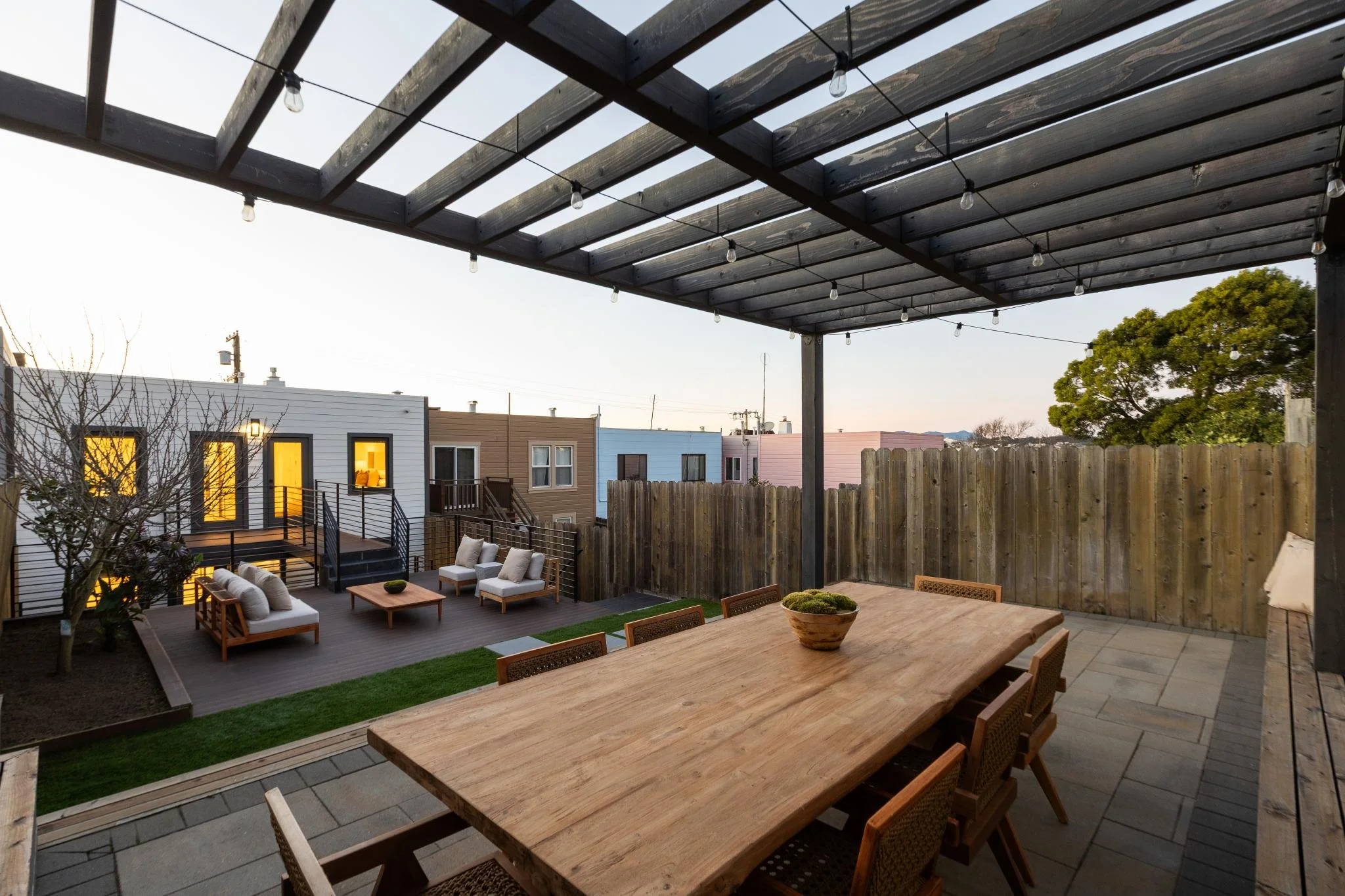 A backyard patio with outdoor seating, a wooden dining table, string lights, and a privacy fence, during dusk.