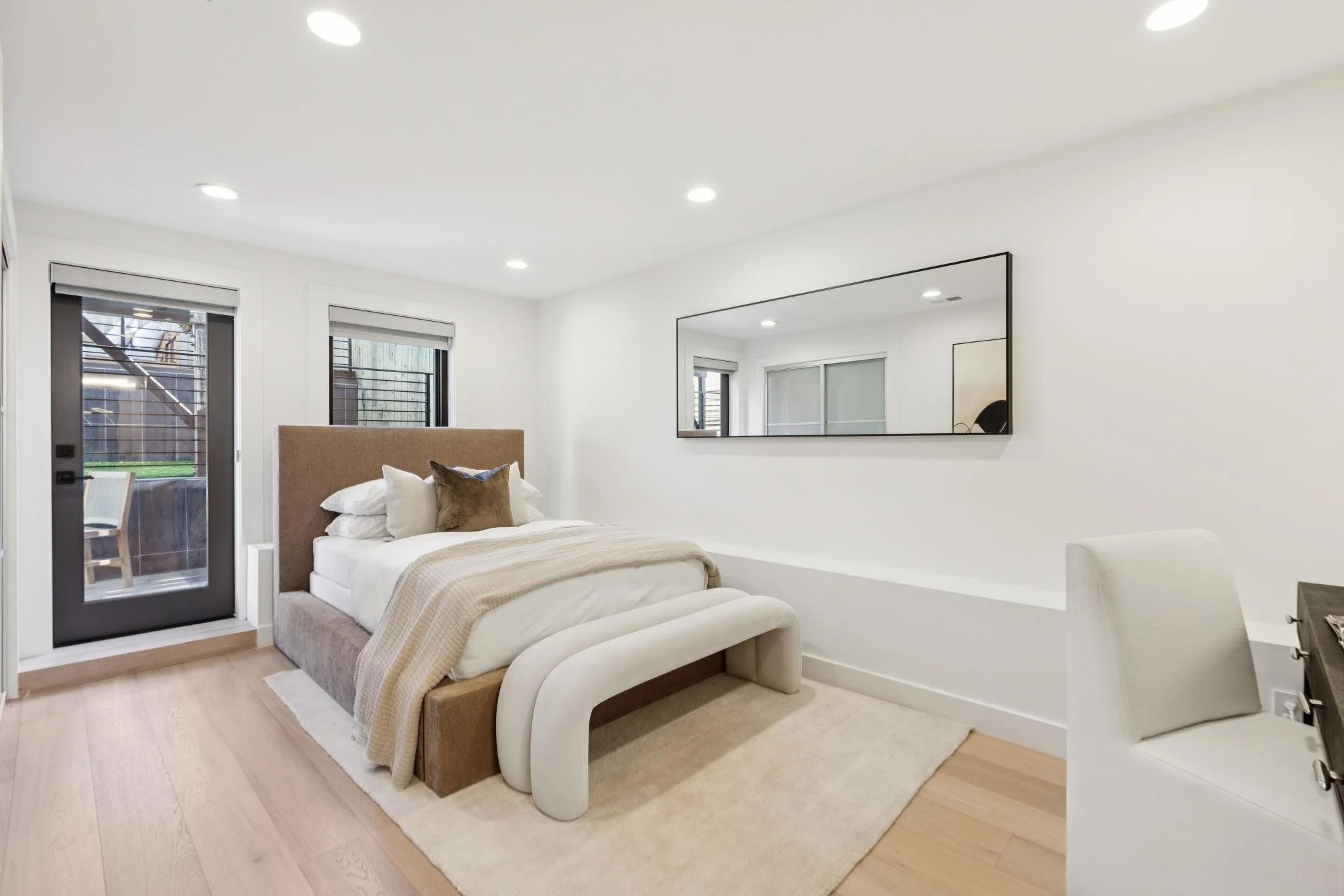 Modern bedroom with beige upholstered bed, white bedding, and brown accent pillow, wooden floor, large wall mirror, and sliding glass door leading to an outdoor patio.
