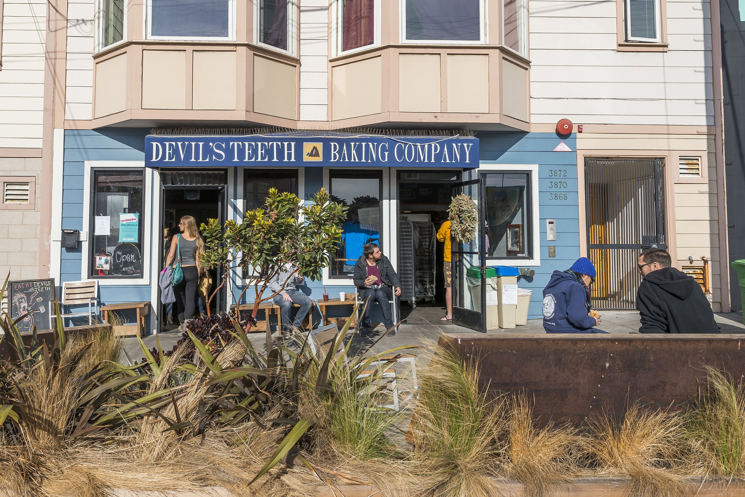 People sitting and standing outside a bakery called 'Devil's Teeth Baking Company' on a sidewalk, with a blue storefront, trees, and plants in the foreground.
