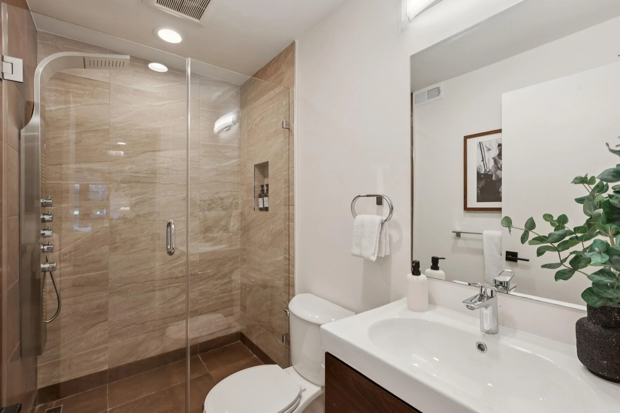 Modern bathroom with a glass-enclosed shower, a white toilet, a sink with a wooden cabinet, a large mirror, and a potted plant. White walls and brown tiled flooring and shower walls.