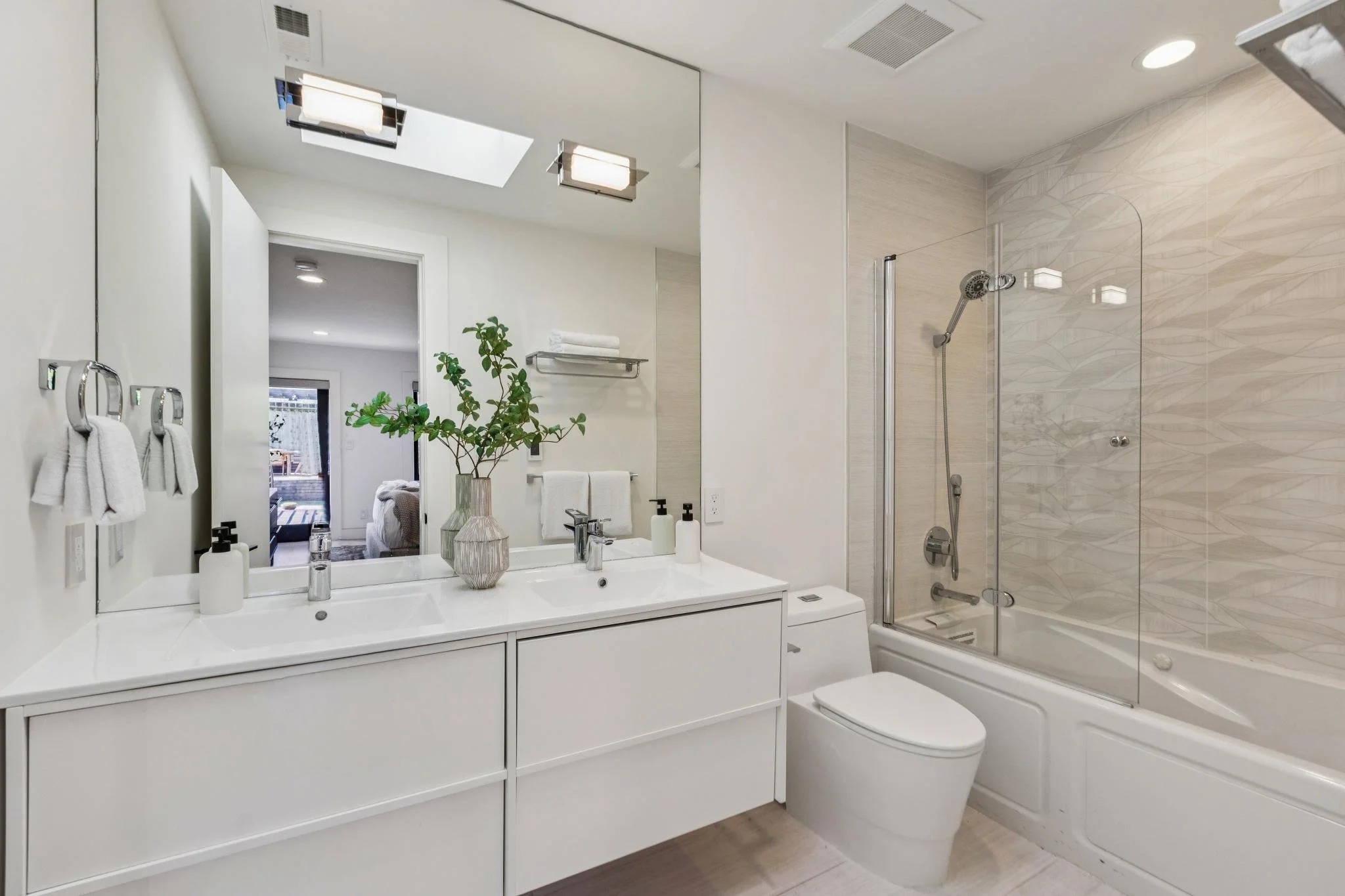 Modern bathroom with double sink vanity, large mirror, potted plant, towels, and a shower bathtub with glass door.