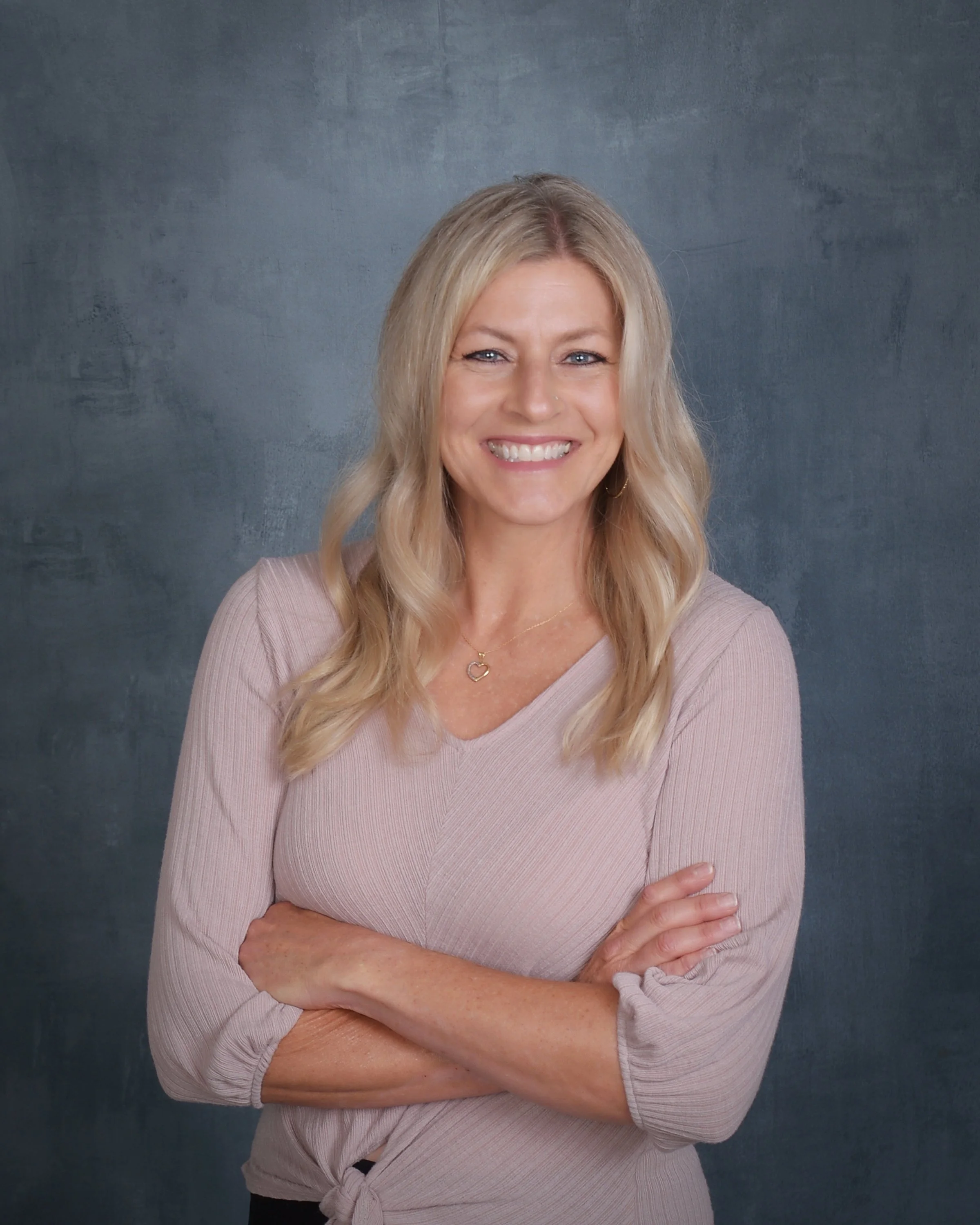 A woman with long blonde hair smiling, crossing her arms, wearing a light pink, long-sleeve top, standing against a textured dark gray background.