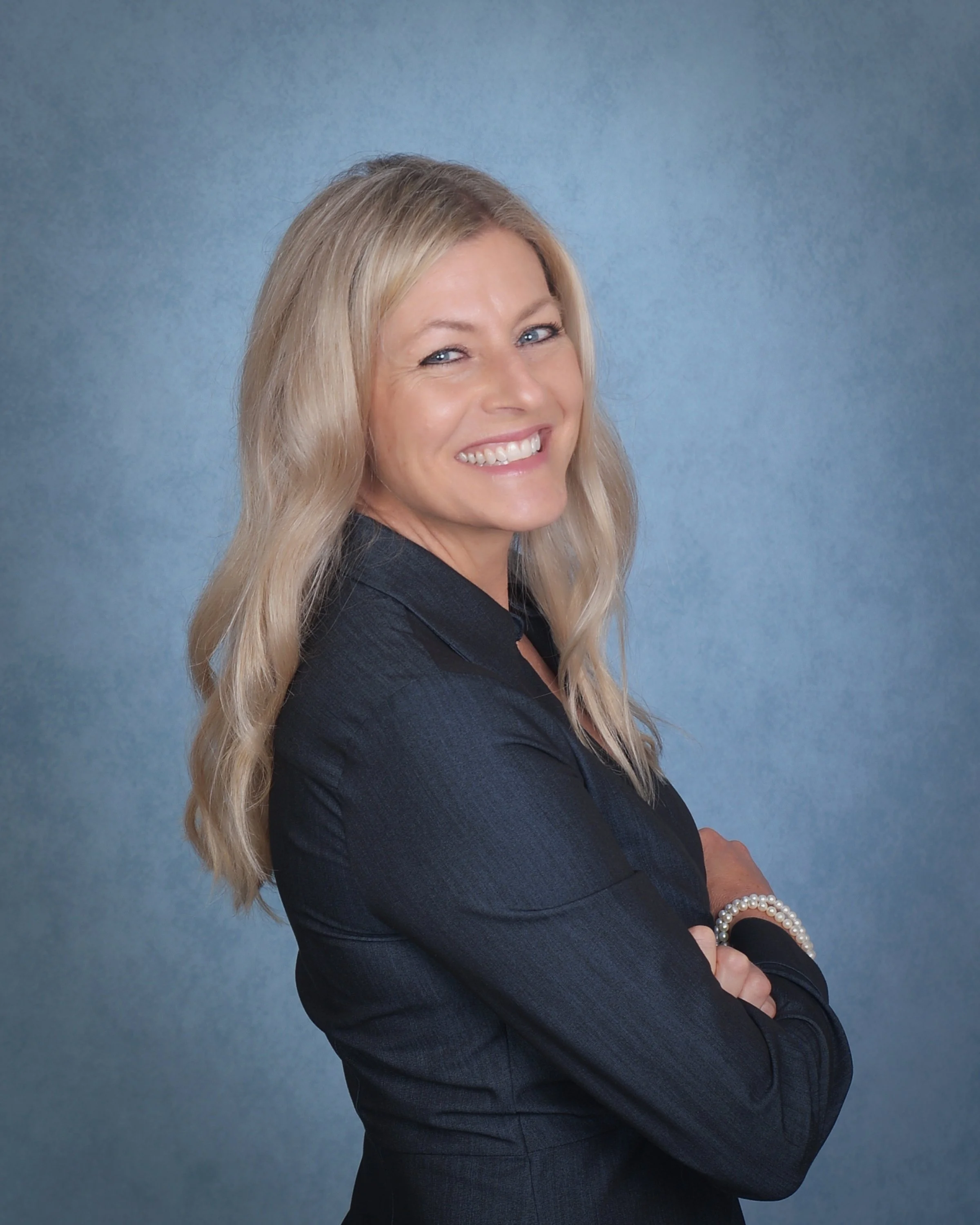 Portrait of a smiling blonde woman with crossed arms, dressed in professional black attire, standing against a blue background.