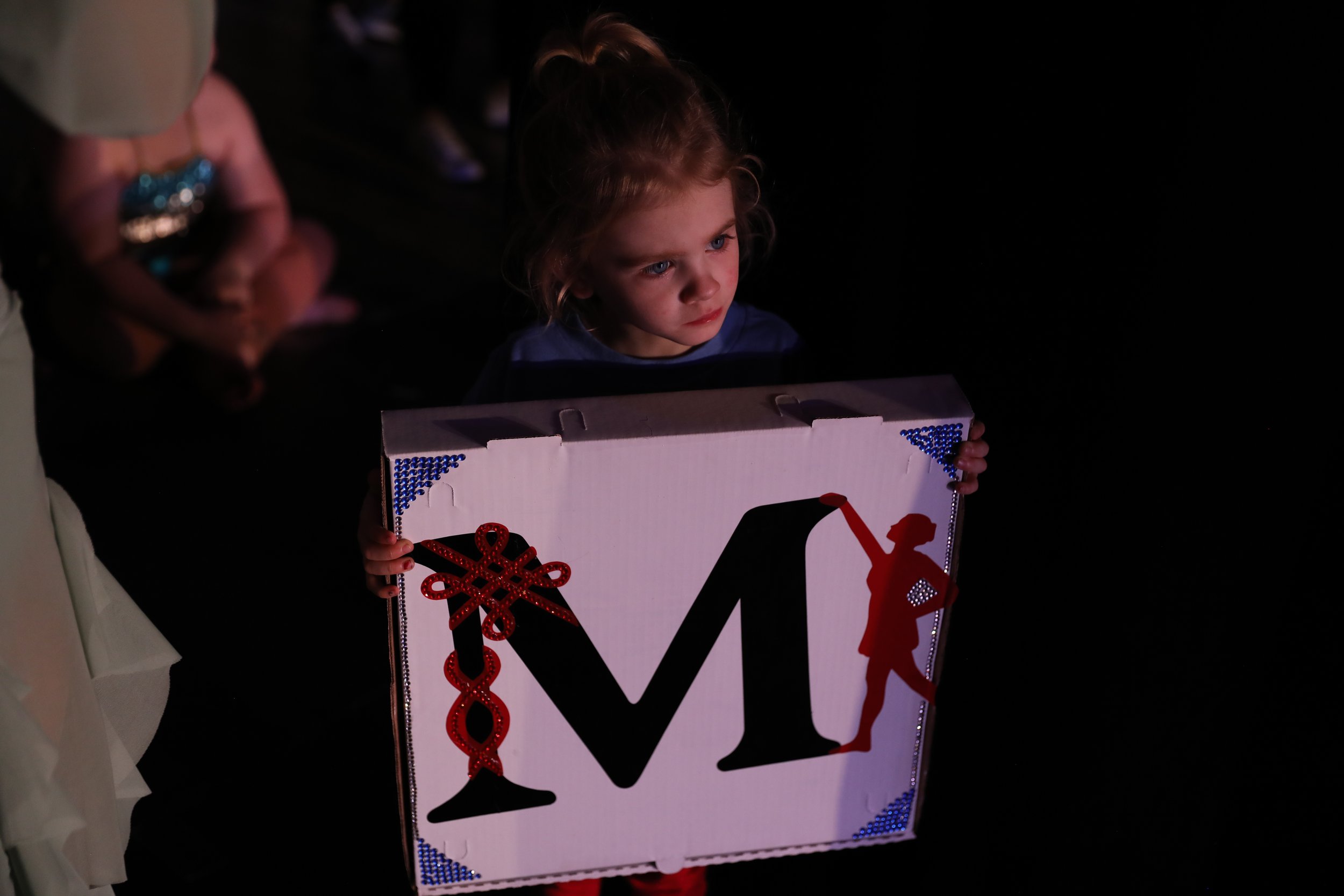 Young girl holding a large box with the letter 'M' and holiday decorations on it, in a dark room.