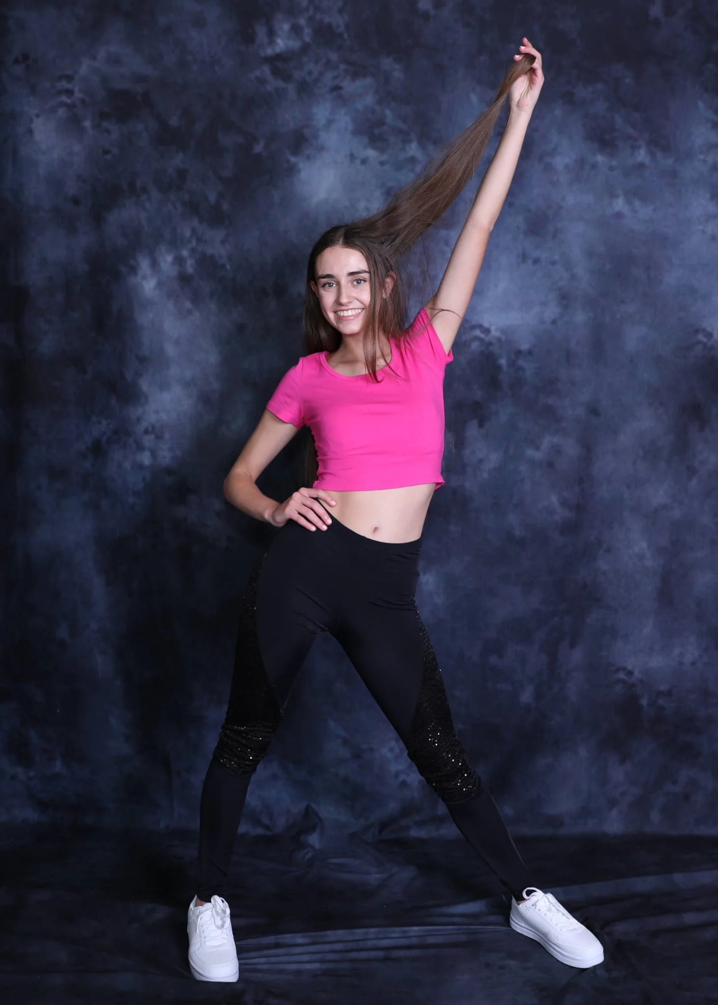 A young woman with long brown hair wearing a pink crop top and black pants, smiling and holding her hair up against a dark, textured background.