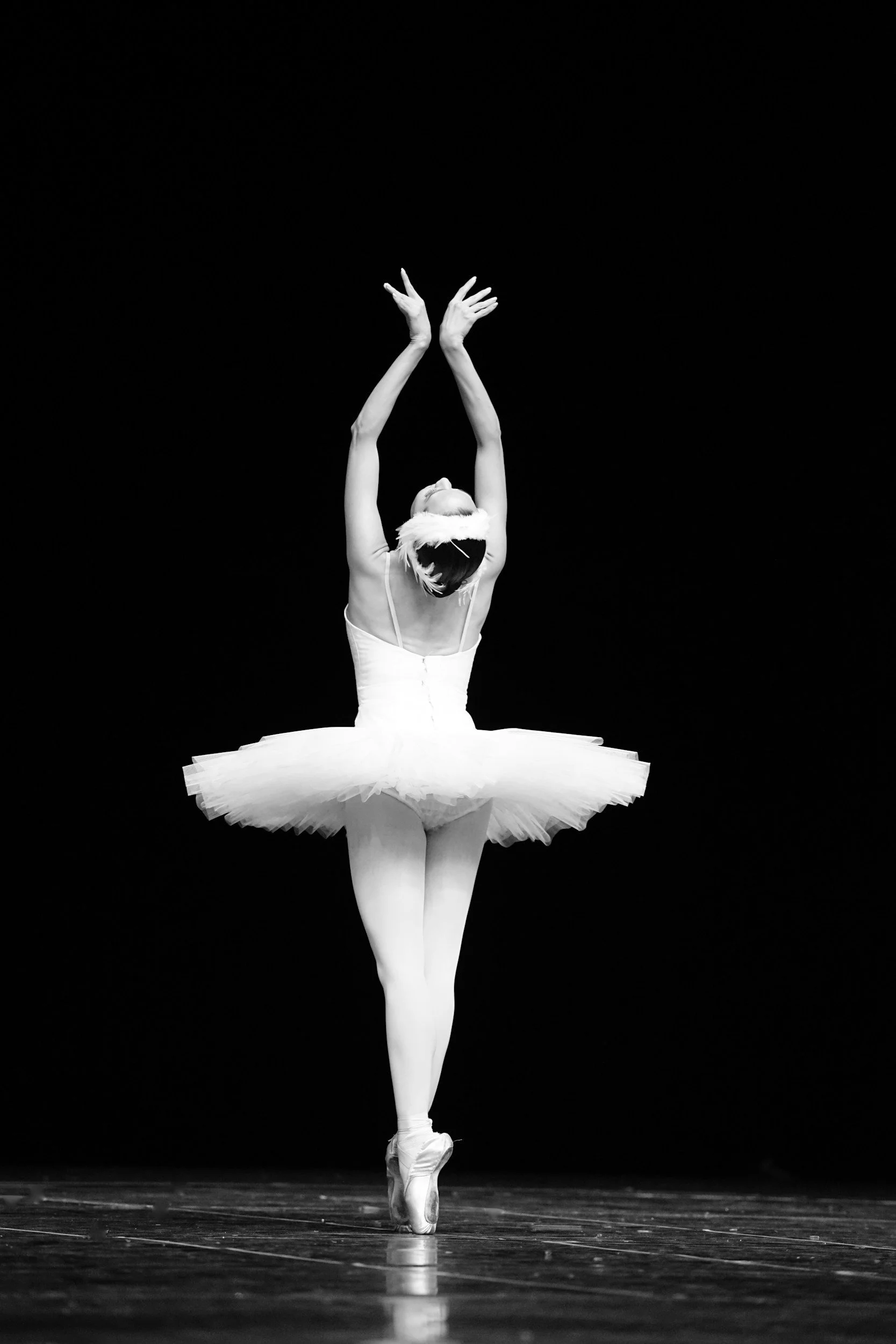 A ballet dancer in a white tutu performing a pose on stage, with arms raised above head and slight bend at the waist, on pointe shoes against a dark background.