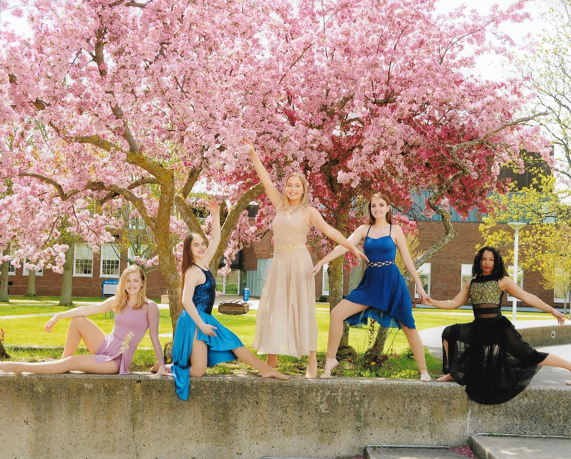 Five women in colorful dresses posing under cherry blossom trees on a college campus.