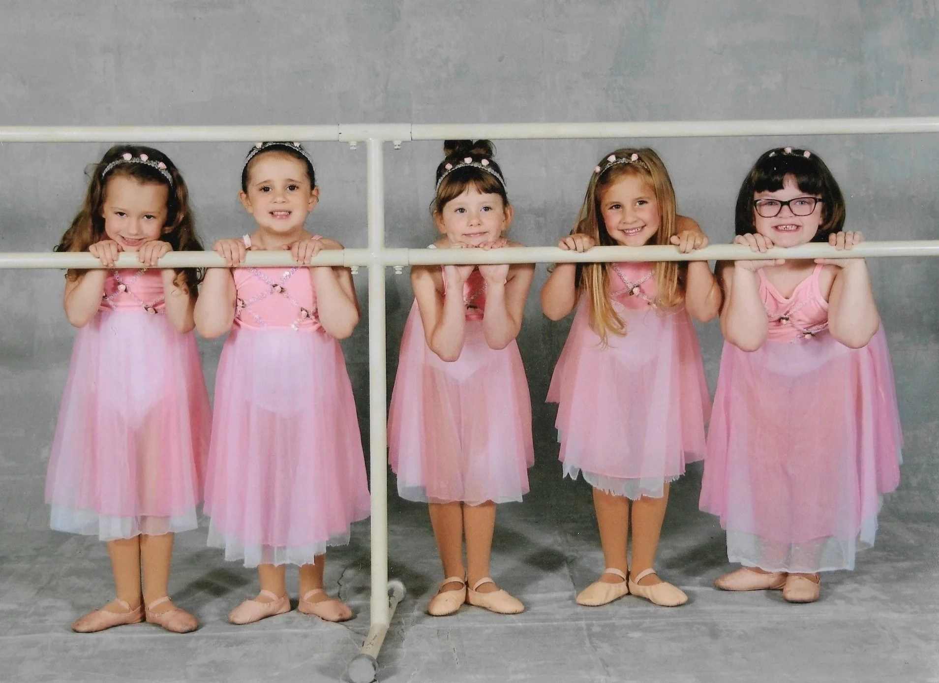 Five young girls in pink ballet dresses standing behind a ballet barre, smiling and holding onto the barre.