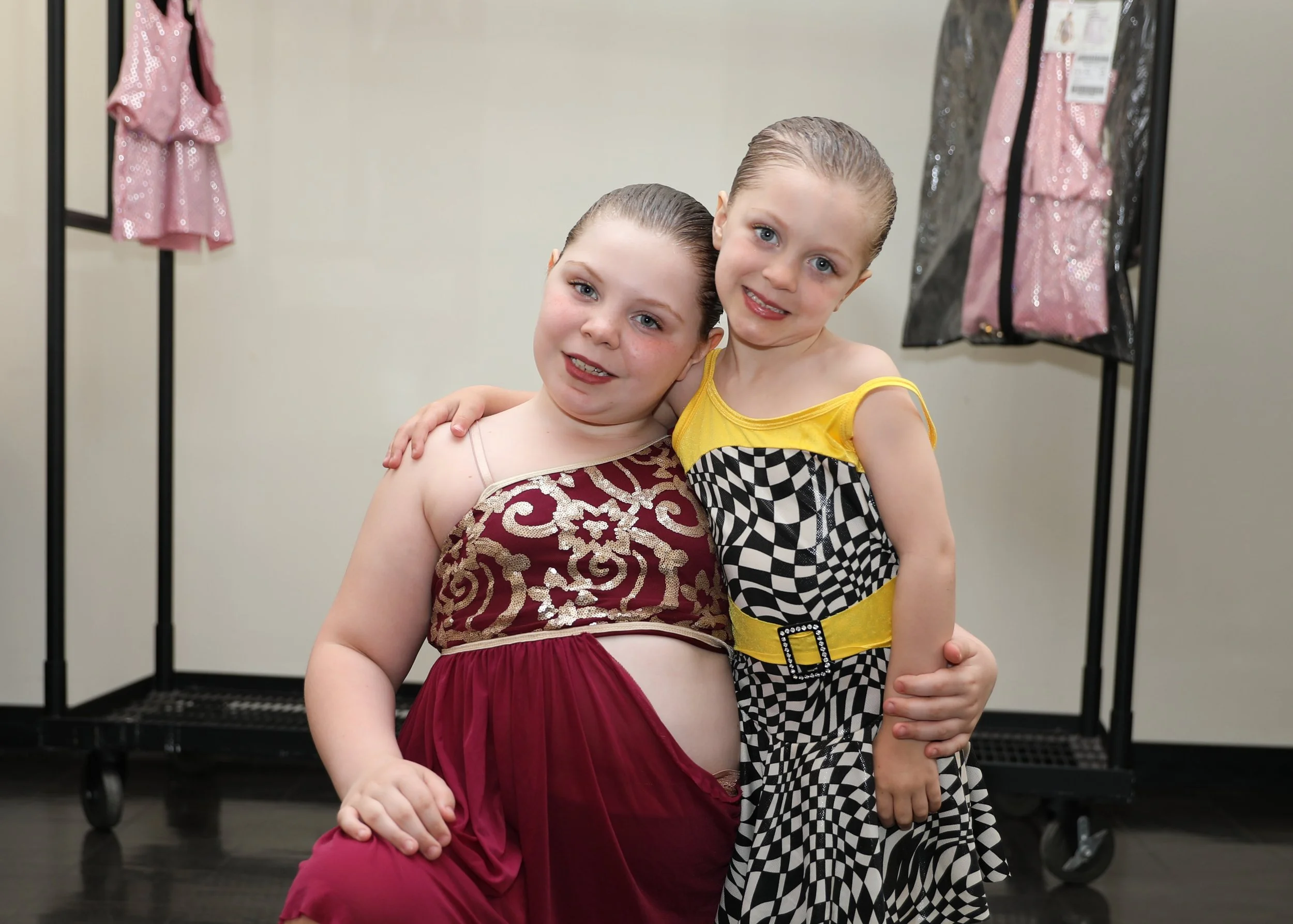 Two young girls with wet hair, smiling and hugging, in a dressing room with clothing on racks in the background.