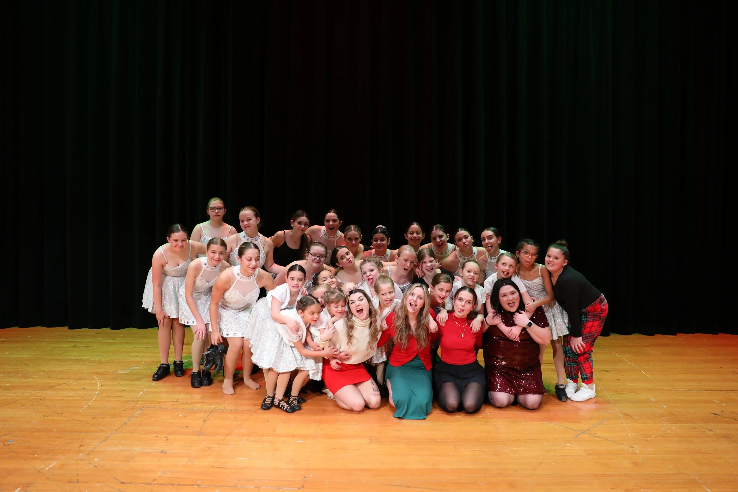 A large group of young girls and women on a wooden stage, posing for a photo in front of black curtains. They are dressed in dance costumes and casual clothing, smiling and making playful gestures.