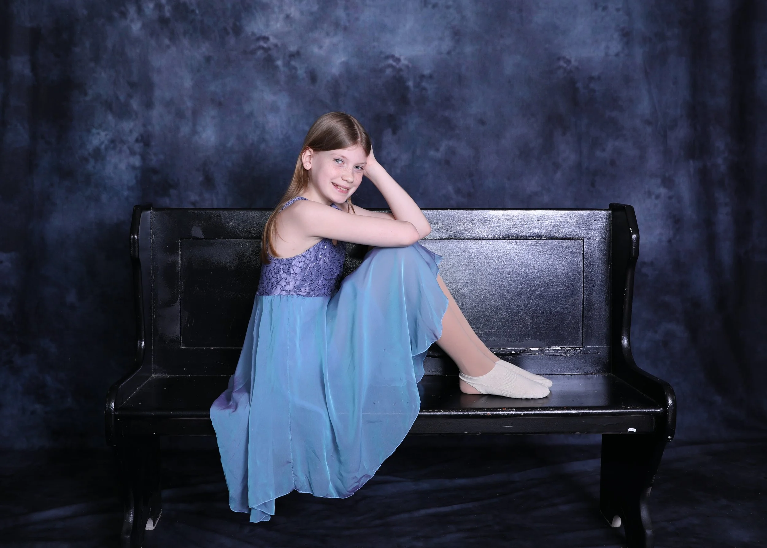 A young girl in a blue dress sitting on a black bench against a dark blue background, smiling with one hand on her head.