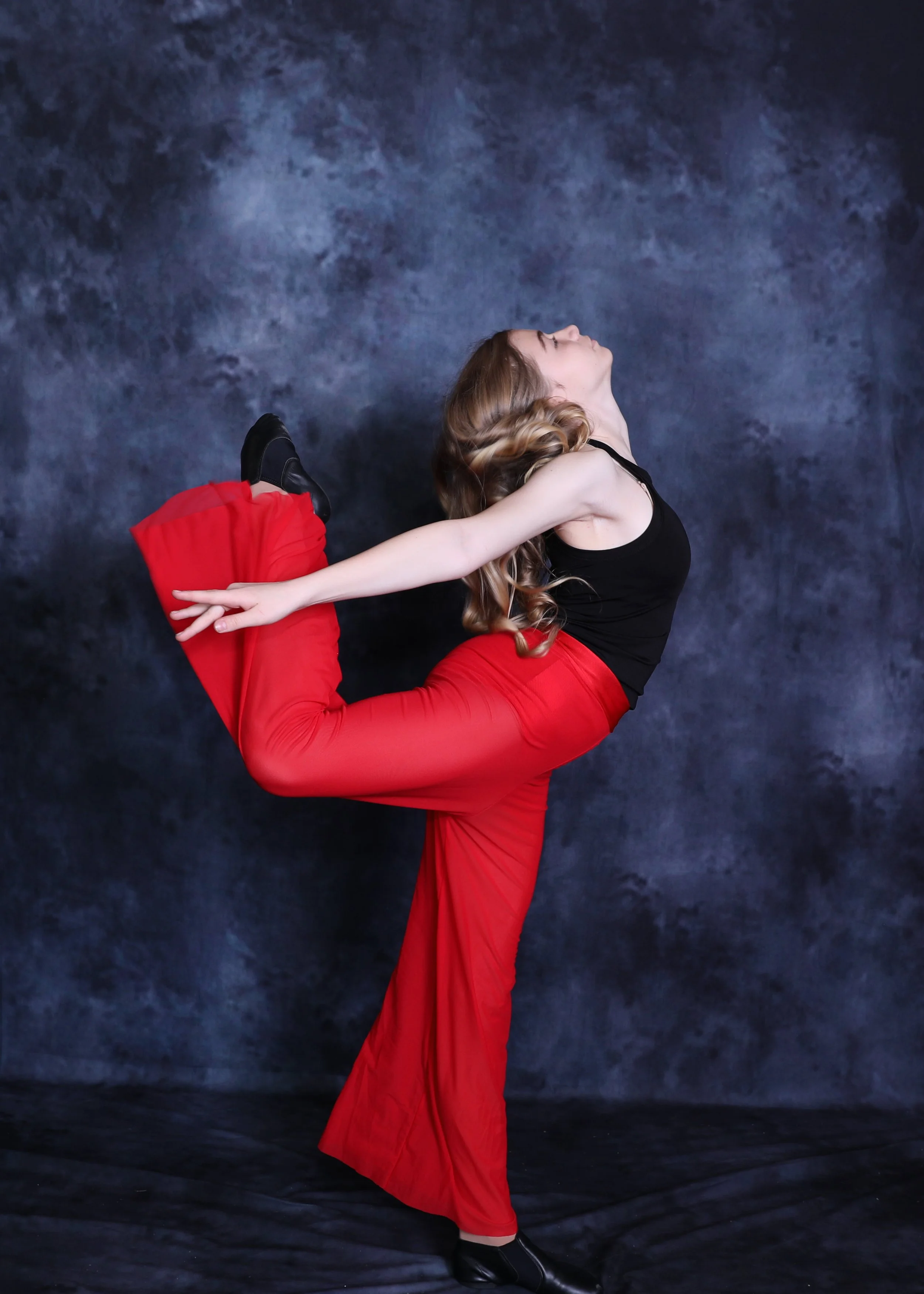 A woman in red pants and a black top performs a dance pose against a dark, textured background, balancing on her right leg with her left leg bent and held behind her, her left arm extended backward, and her head tilted upward.