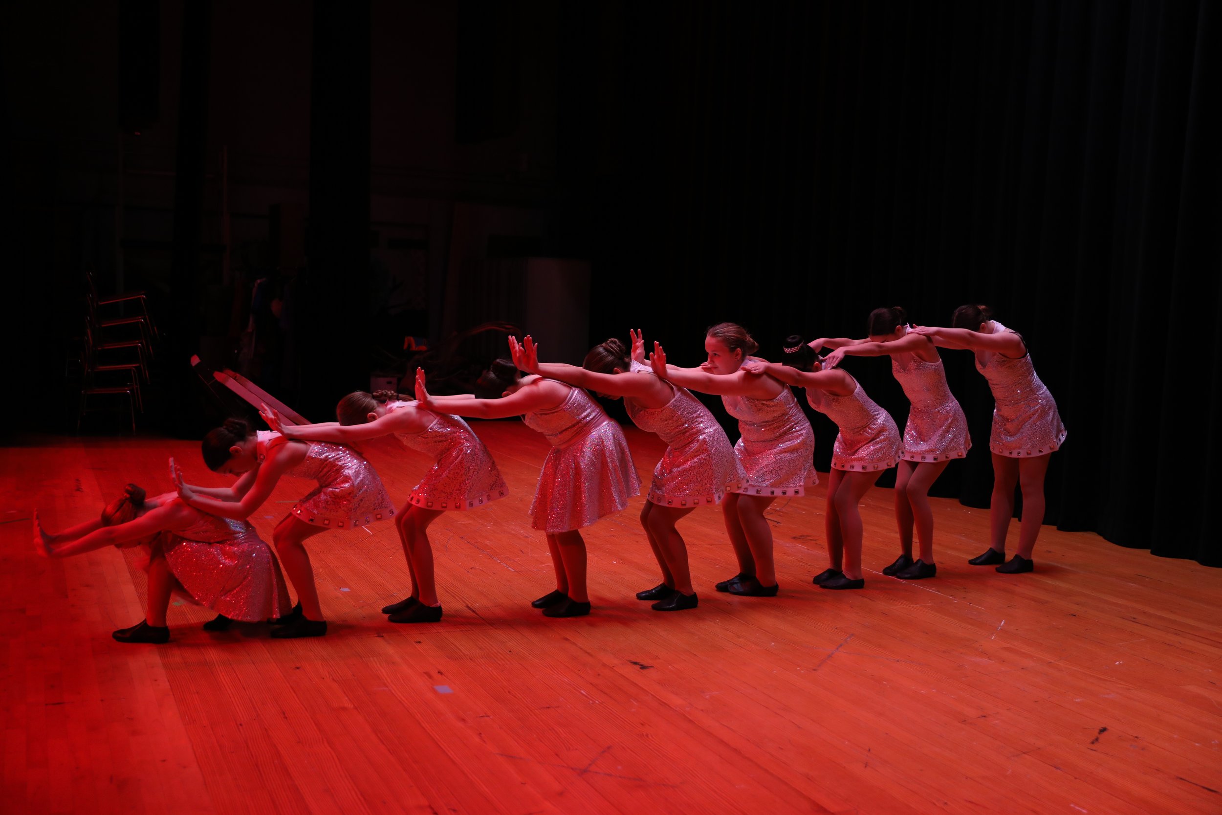 A group of young girls in shiny pink dresses performing a dance on a wooden stage with black curtains.
