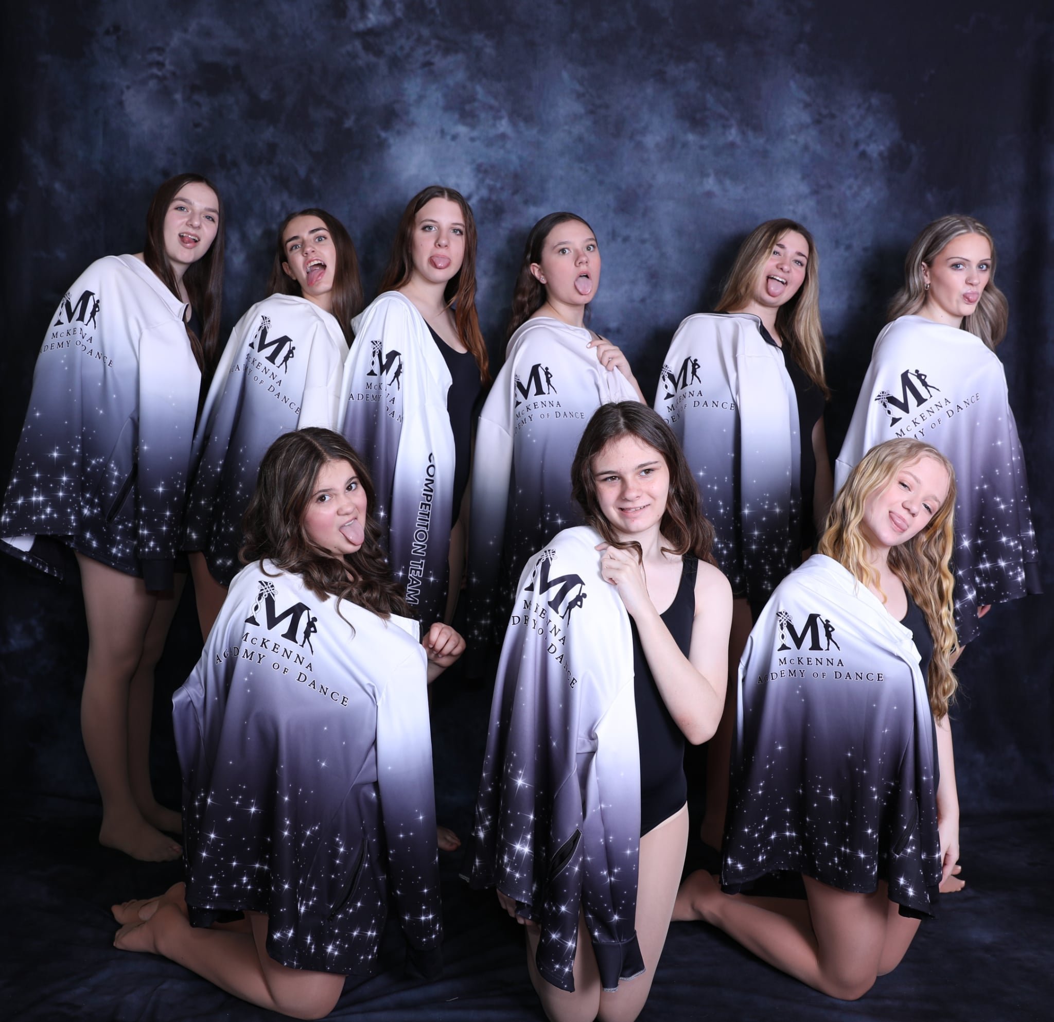 Group of young female dancers posing in dance team hoodies, making silly faces, in front of a dark mottled backdrop.