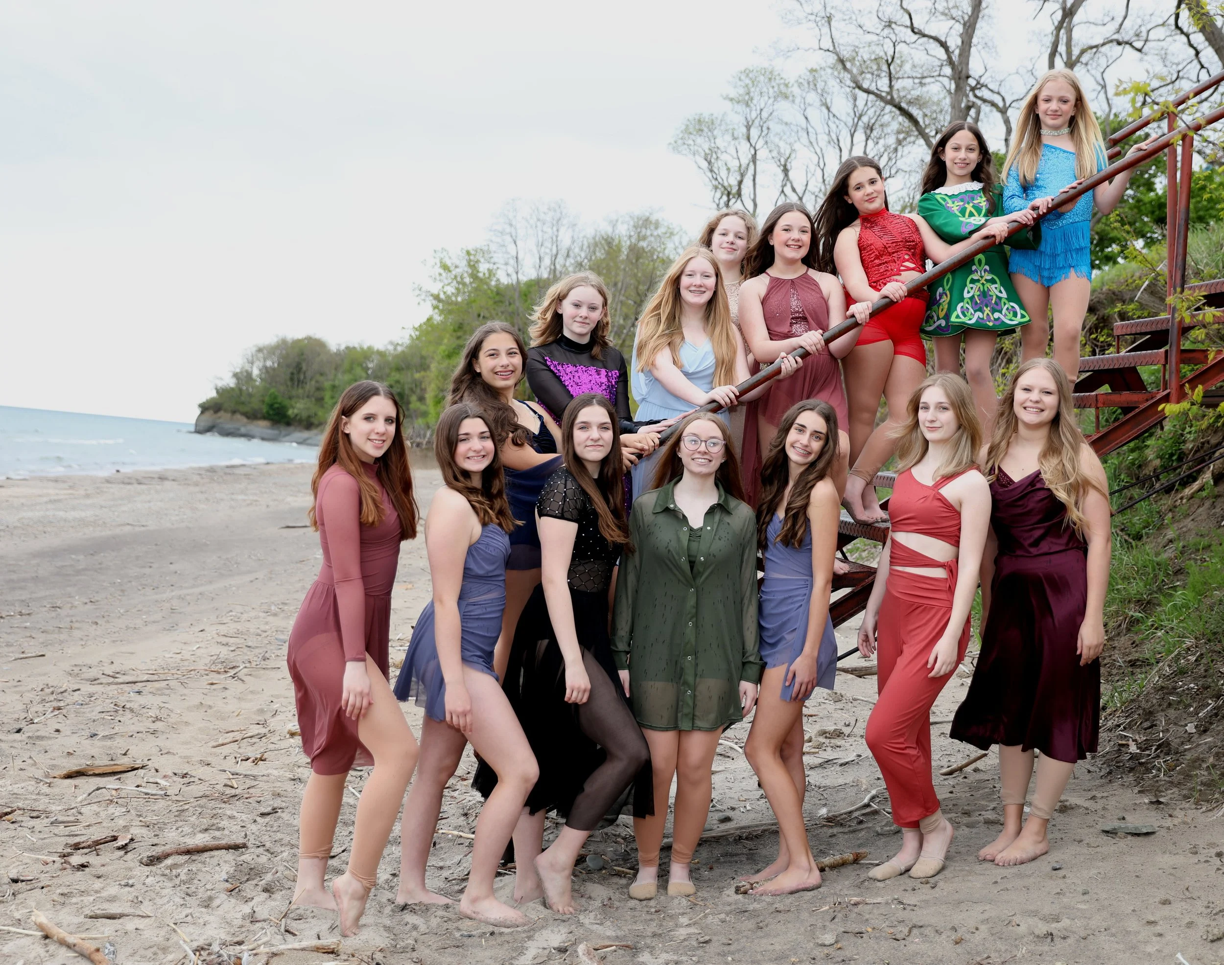 Group of teenage girls on a beach, some standing and some on stairs, dressed in colorful dresses and posing for a photo.