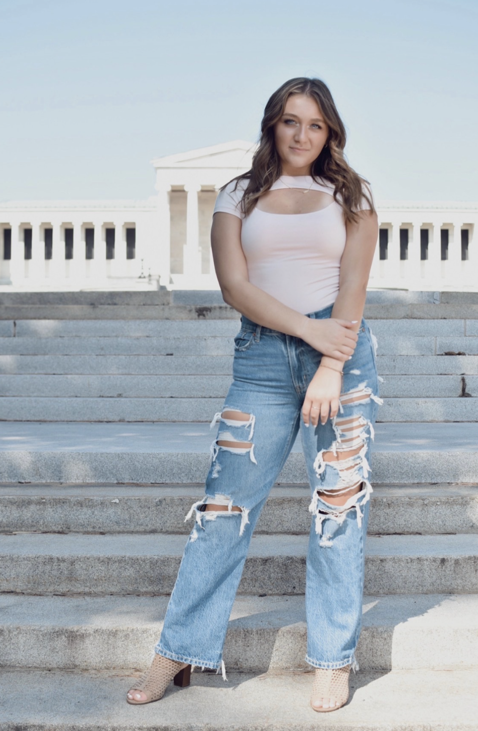 Young woman standing on steps outside the Lincoln Memorial in Washington, D.C., wearing a white top with cutouts, ripped jeans, and open-toed heels.