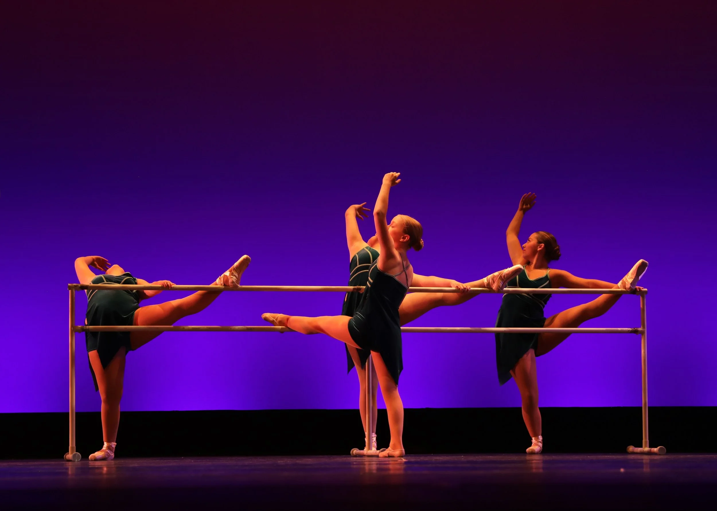 Ballet dancers practicing at a barre on stage with purple background