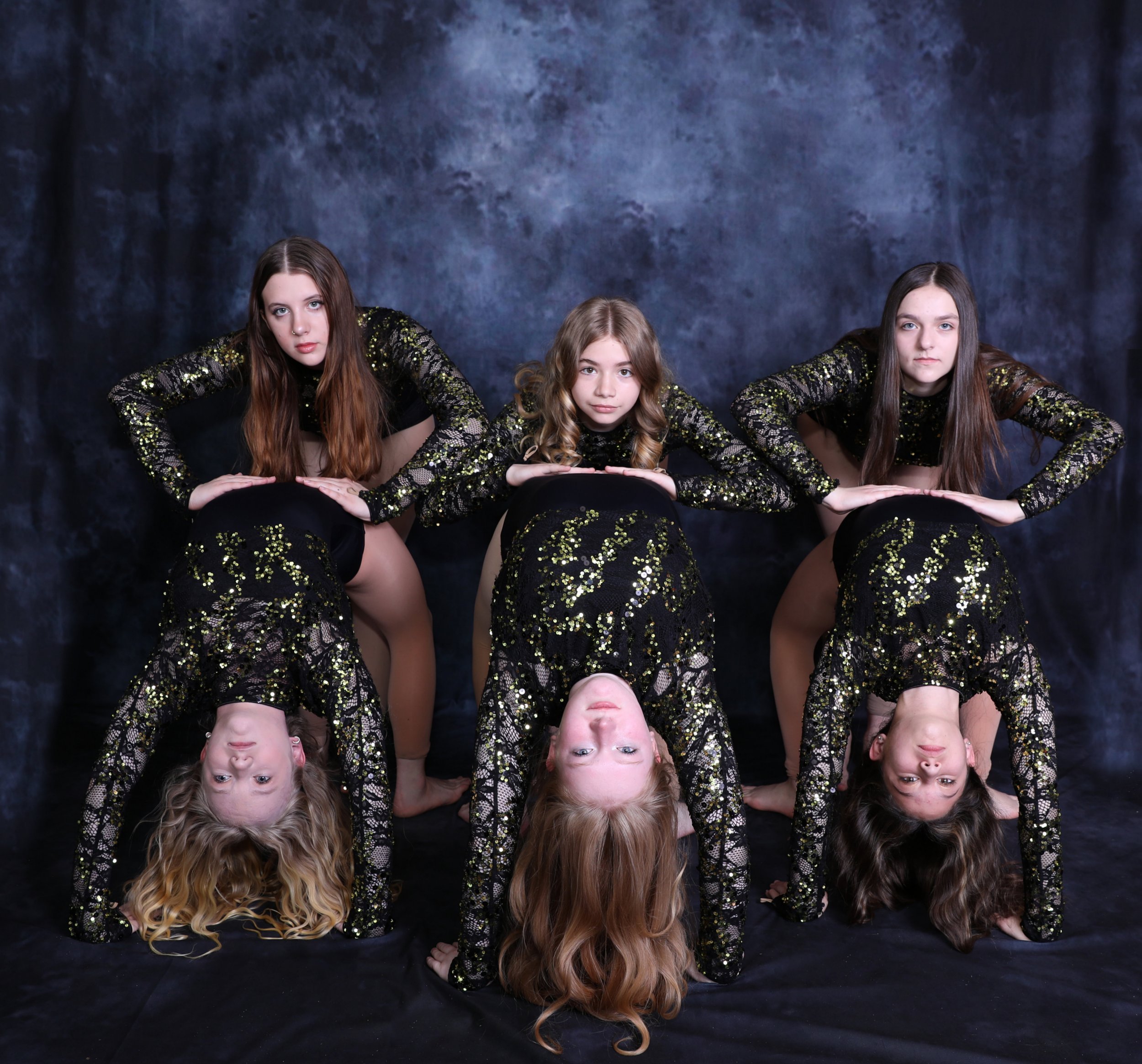 Six young girls with long hair in black and gold sequined costumes perform a handstand pose against a dark backdrop, with three girls upright and three upside down.