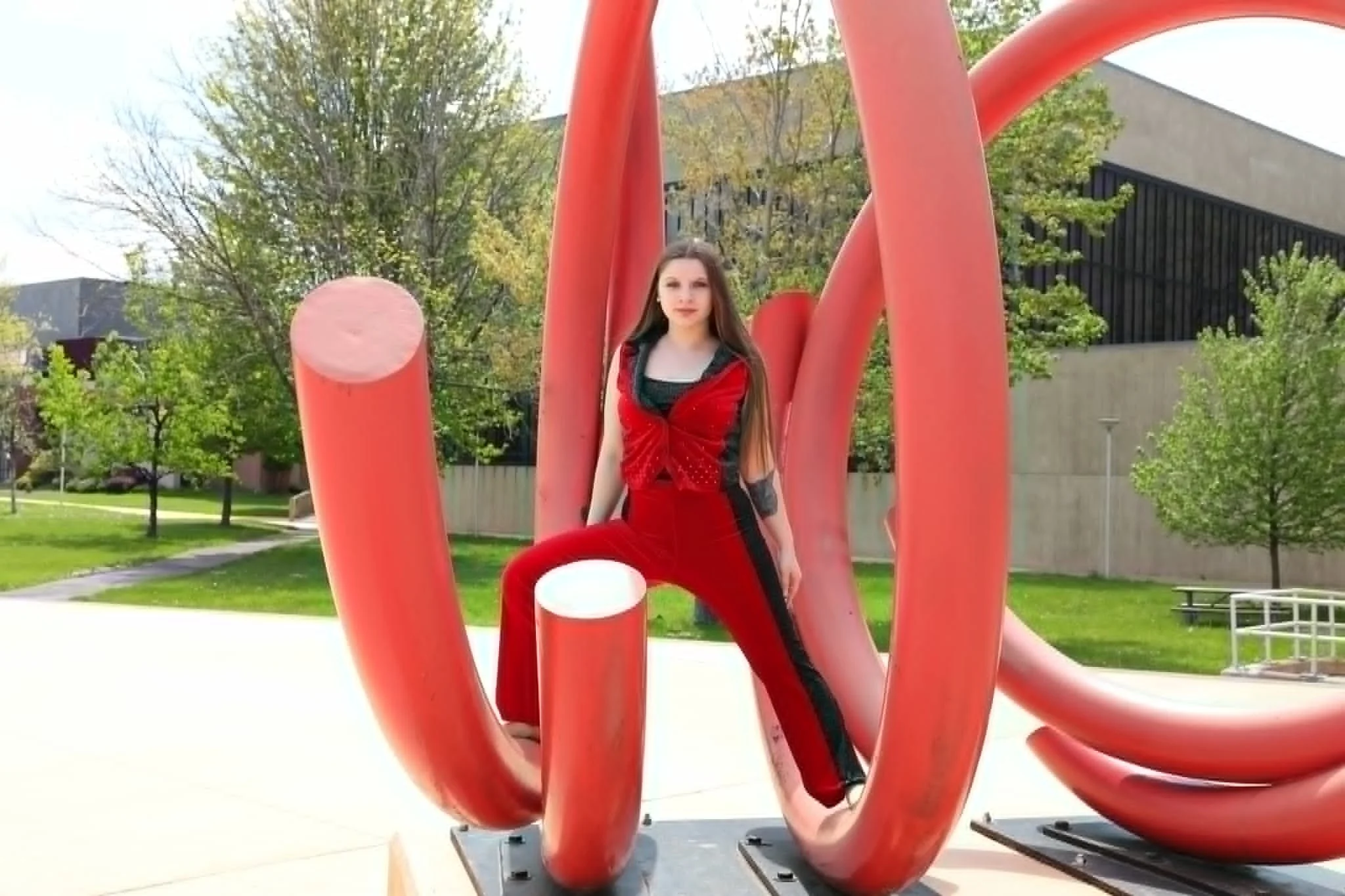 A young woman in a red and black outfit posing on red abstract outdoor sculptures in a park with trees and a modern building in the background.