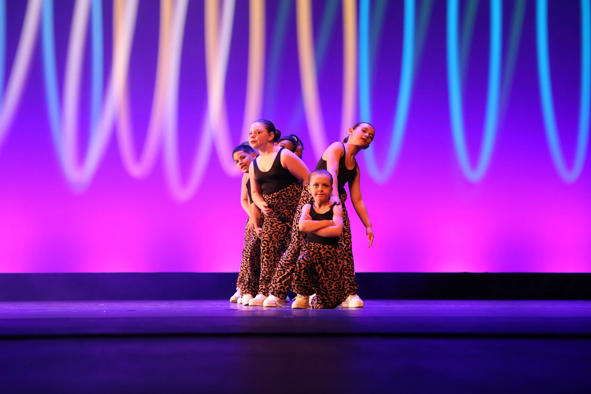 Group of young girls performing a dance on stage, wearing matching leopard print pants and black tops, with purple stage lighting in the background.