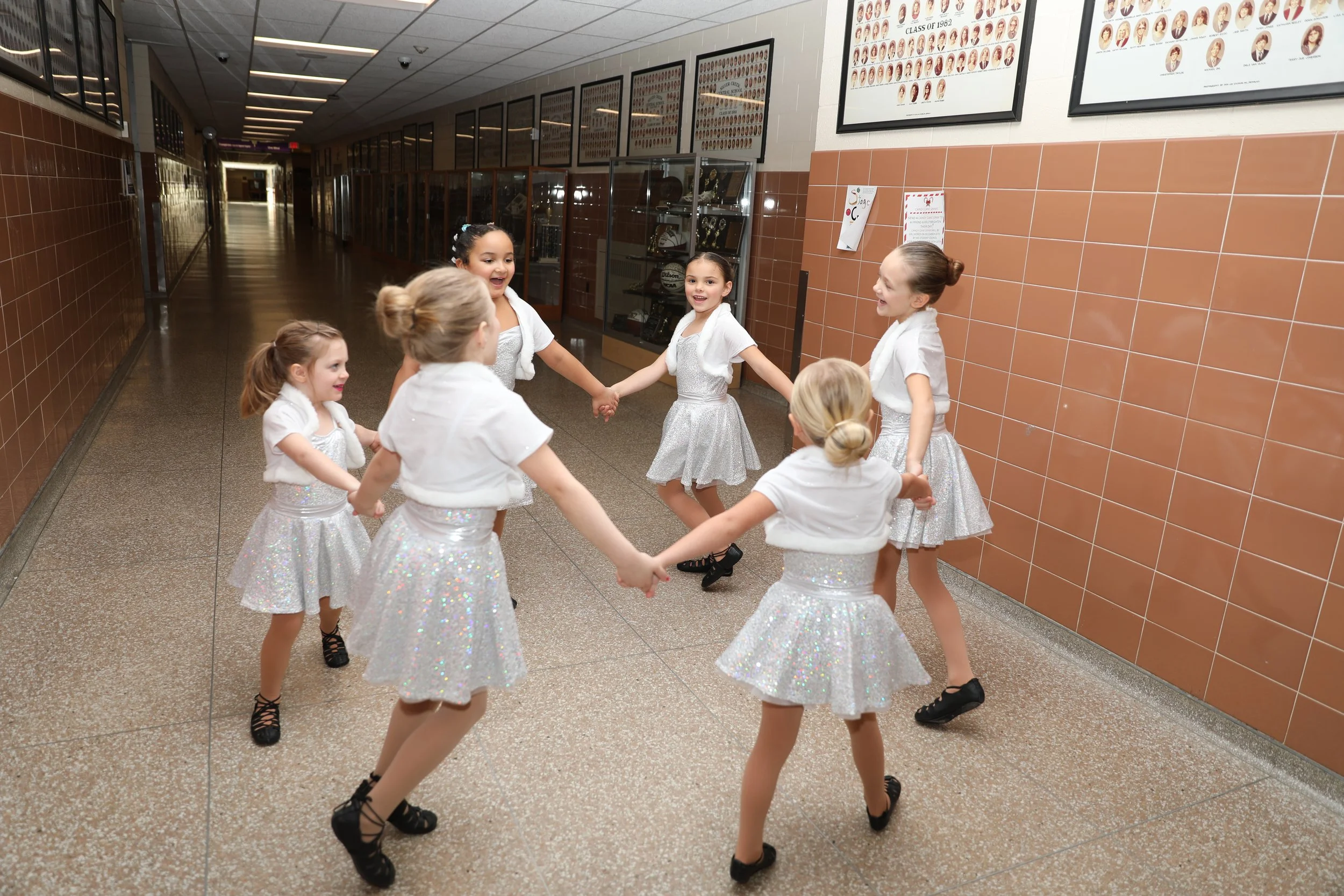 Young girls in sparkly white costumes holding hands and dancing in a school hallway.
