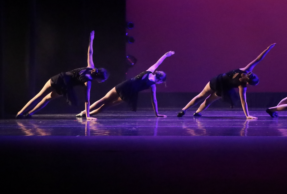 Four female dancers in black costumes performing a dance routine on stage with purple lighting.