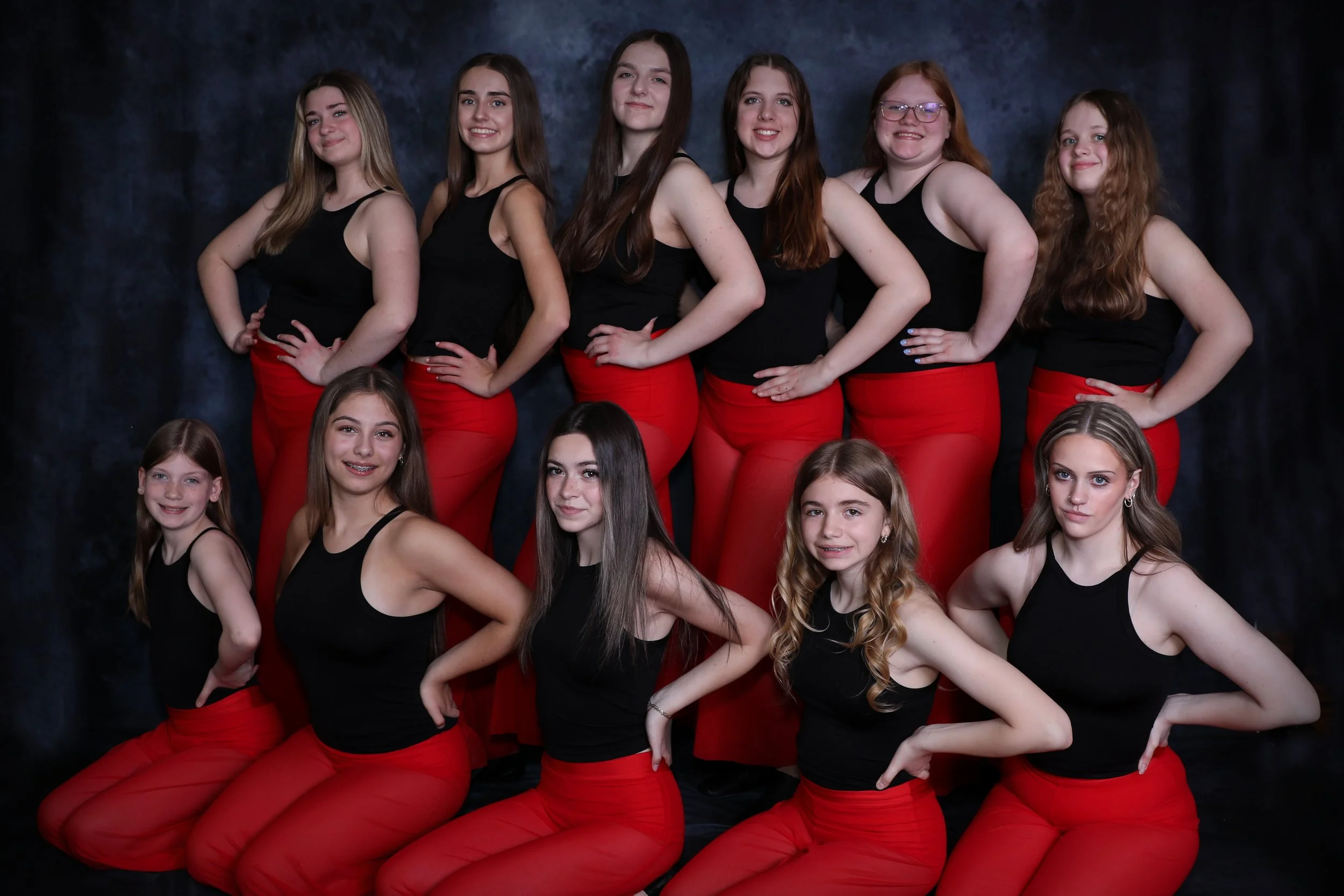 Group of ten girls in matching black tank tops and red pants posing for a team photo against a dark background. They are arranged in two rows, with some standing and some kneeling.