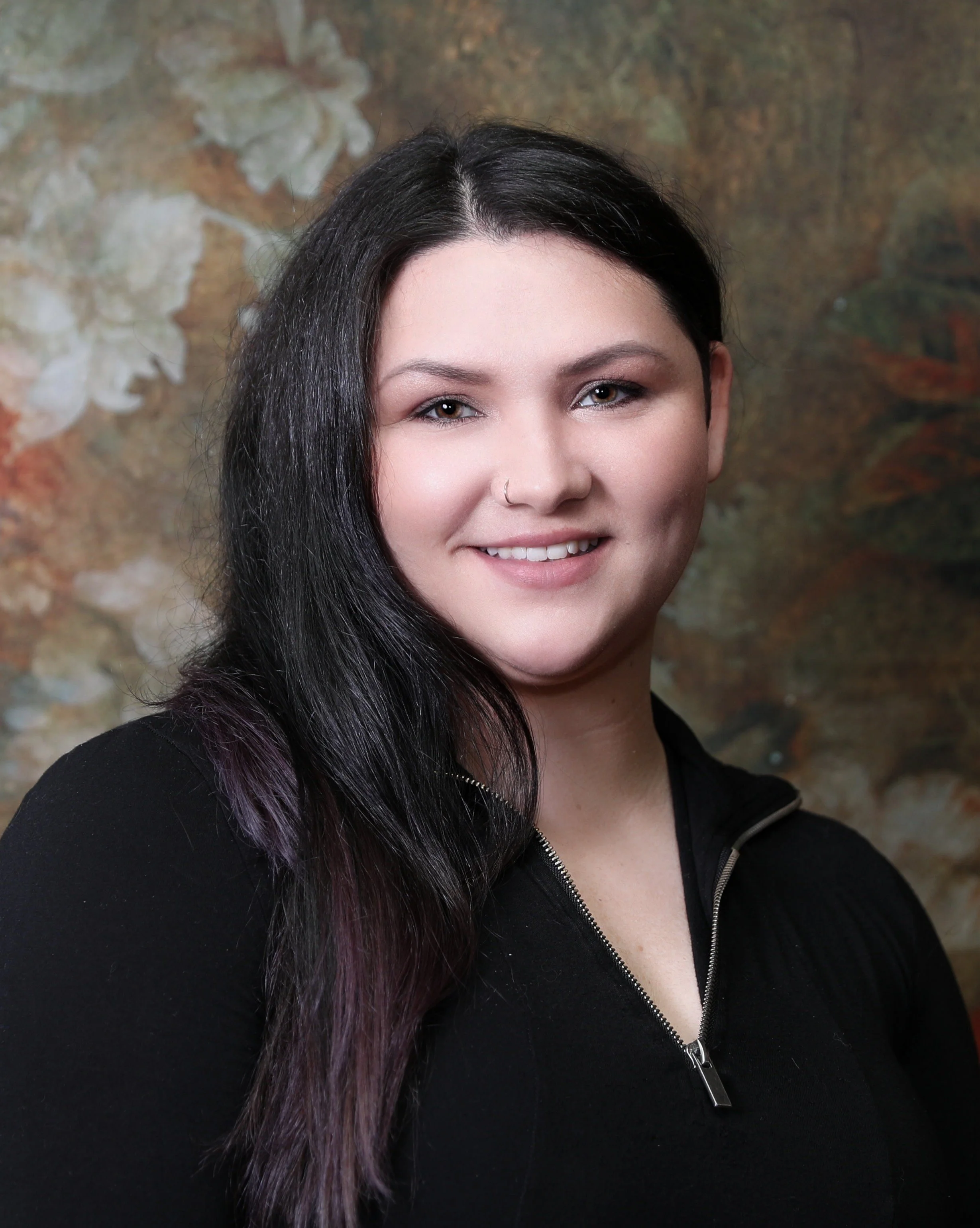 Close-up portrait of a smiling woman with long dark hair and a nose piercing, wearing a black zip-up top against a blurred, textured background. Dance studio owner who is kind and caring.