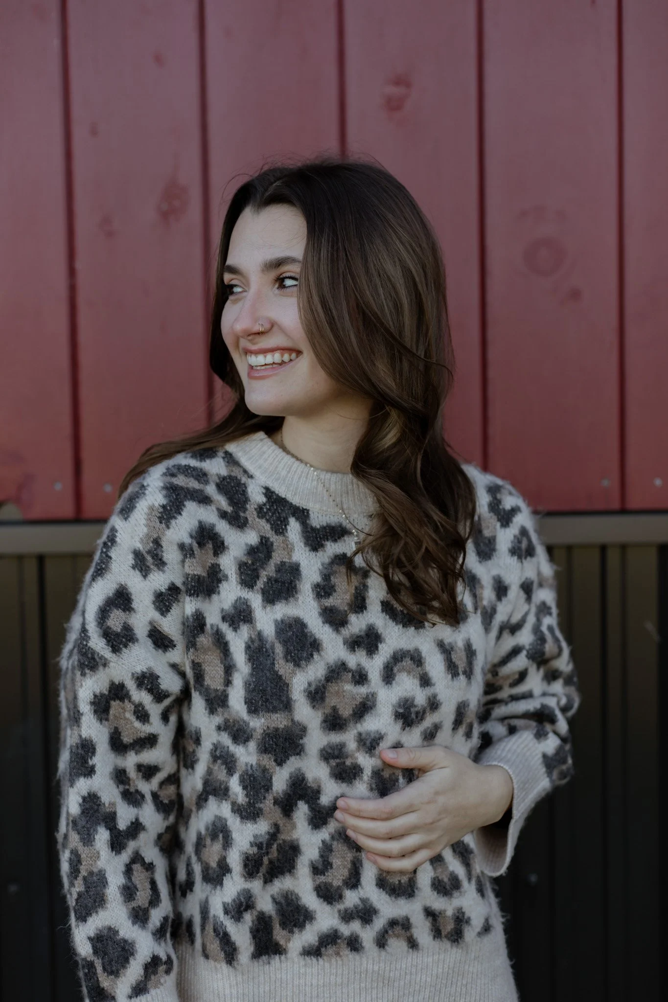 A young woman with long, wavy brown hair smiling and looking to her left, standing in front of a red wooden fence. She is wearing a leopard-print sweater.