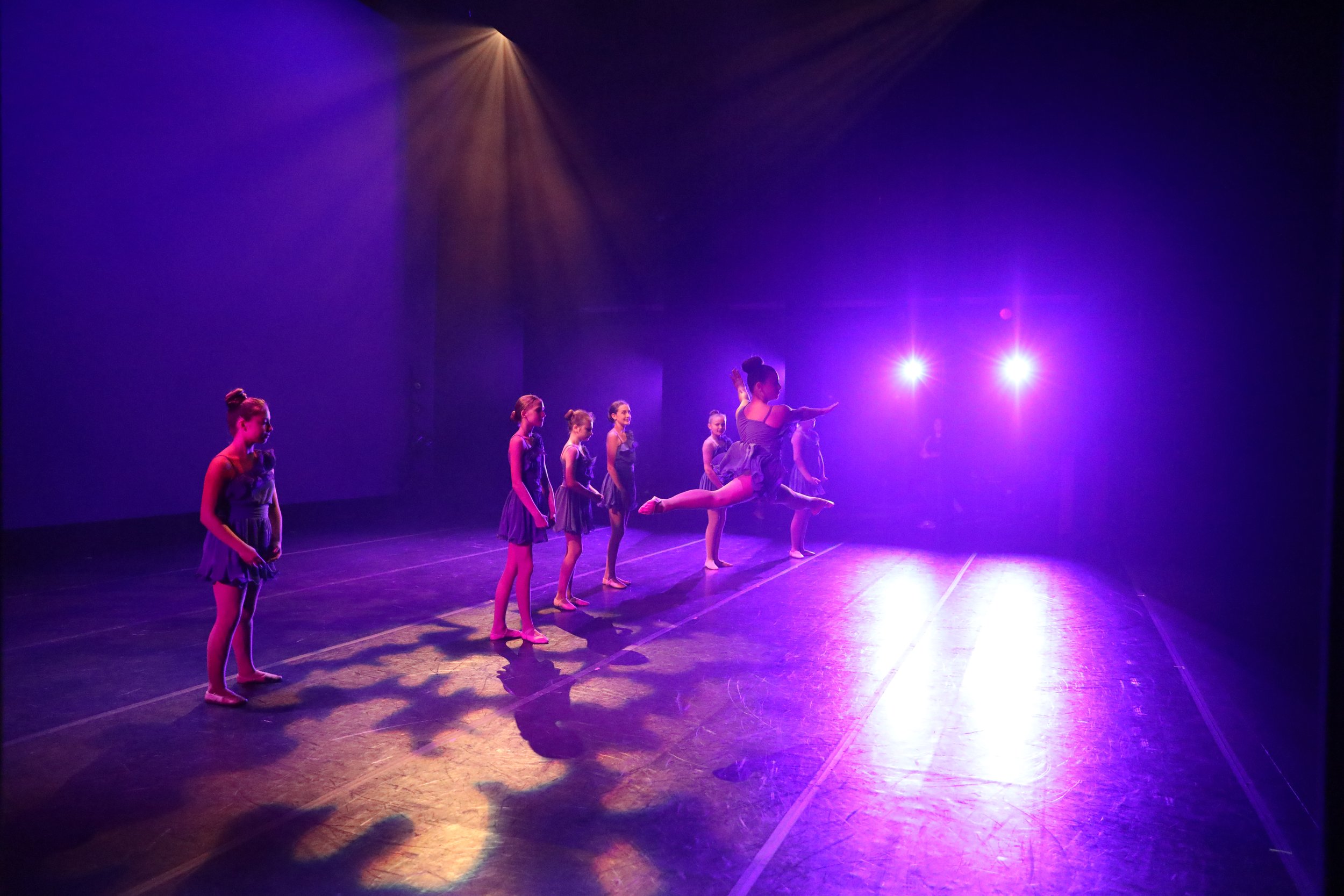 A group of young ballet dancers practicing on stage under purple and yellow stage lights, with one dancer in mid-air doing a leap.