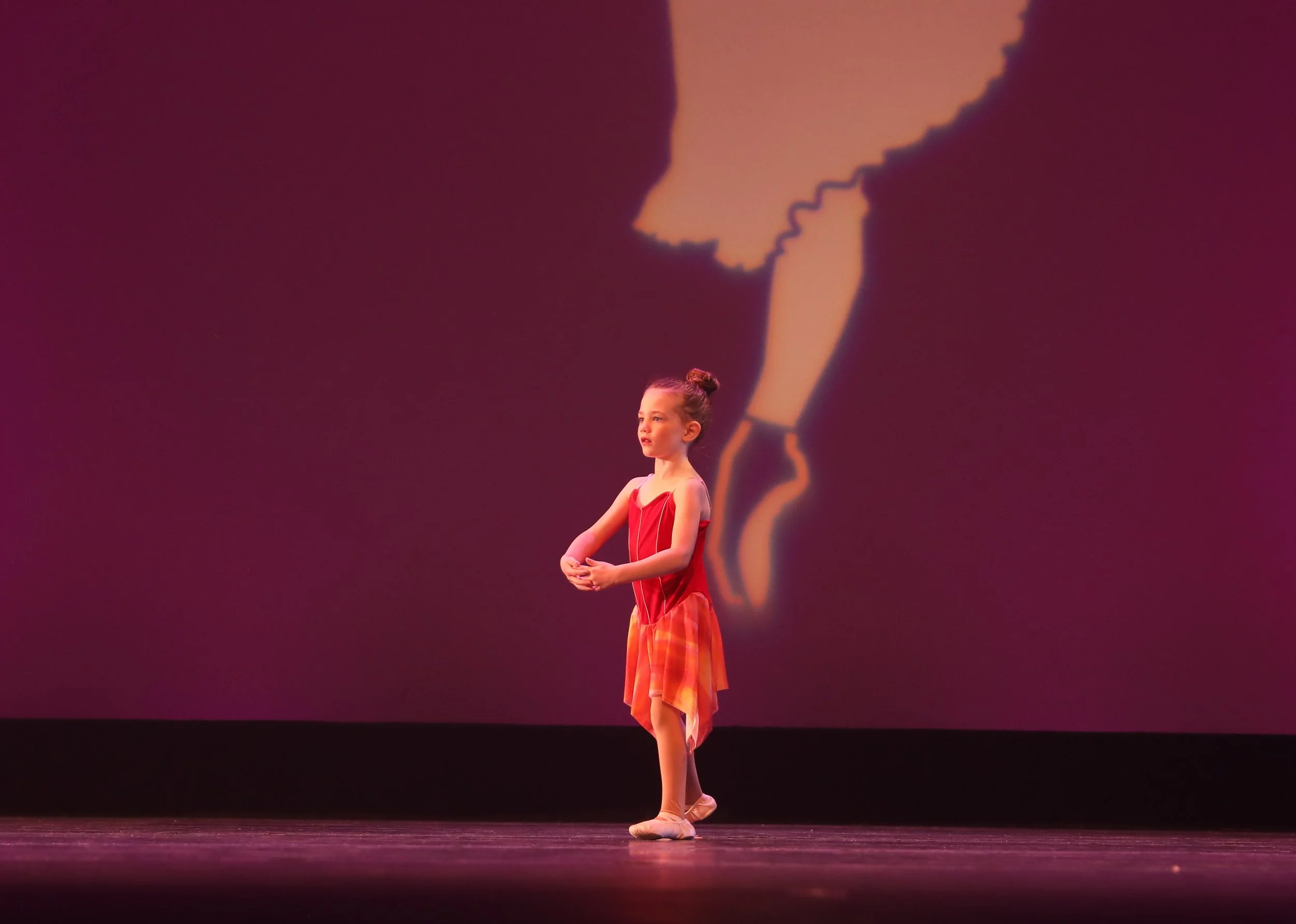 A young girl in a red dress and ballet shoes performing a dance on stage with a projection of a woman in a red dress behind her.