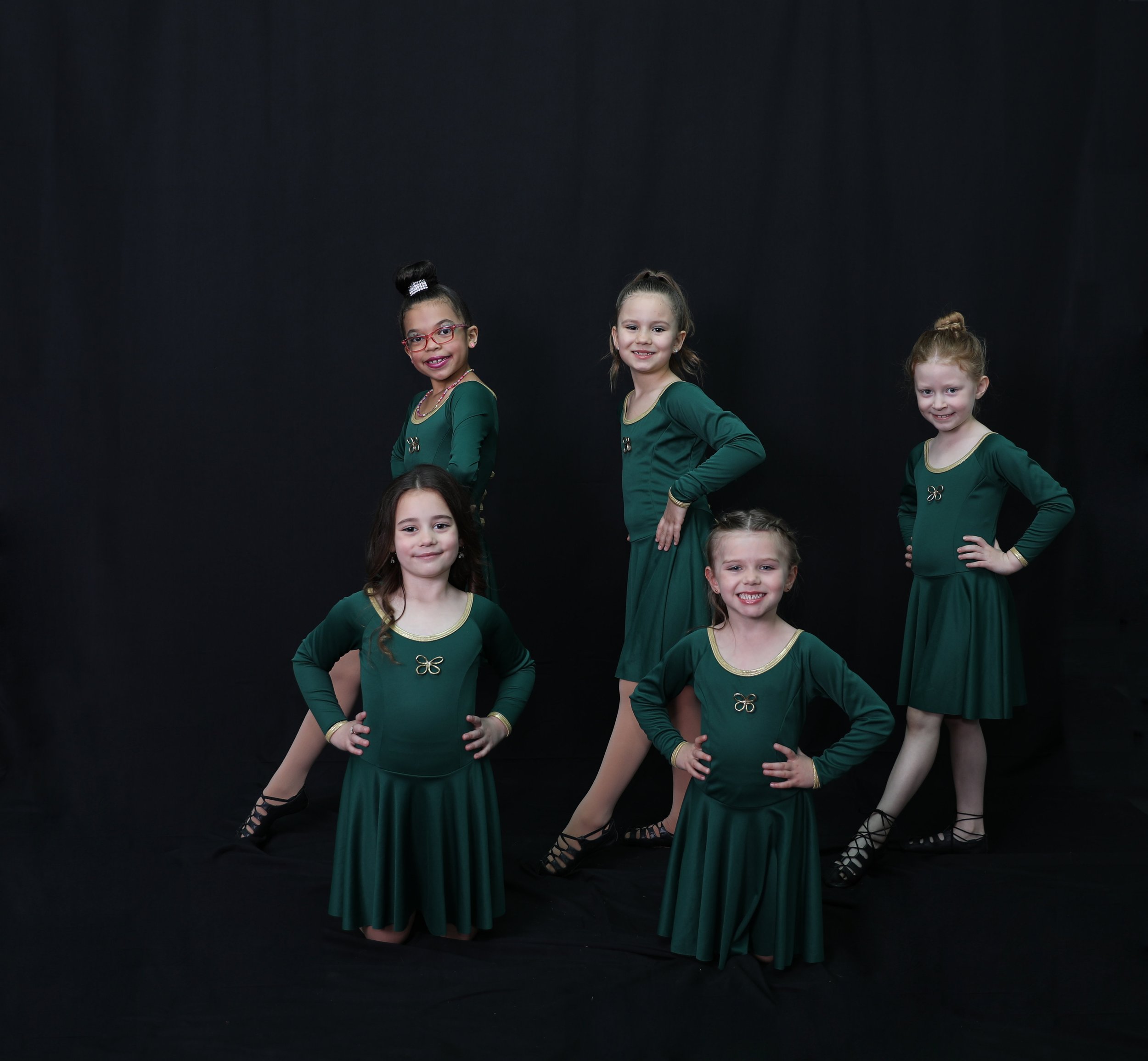 Five young girls in matching dark green dresses with gold accents, posing confidently against a black background, ready for a dance performance.