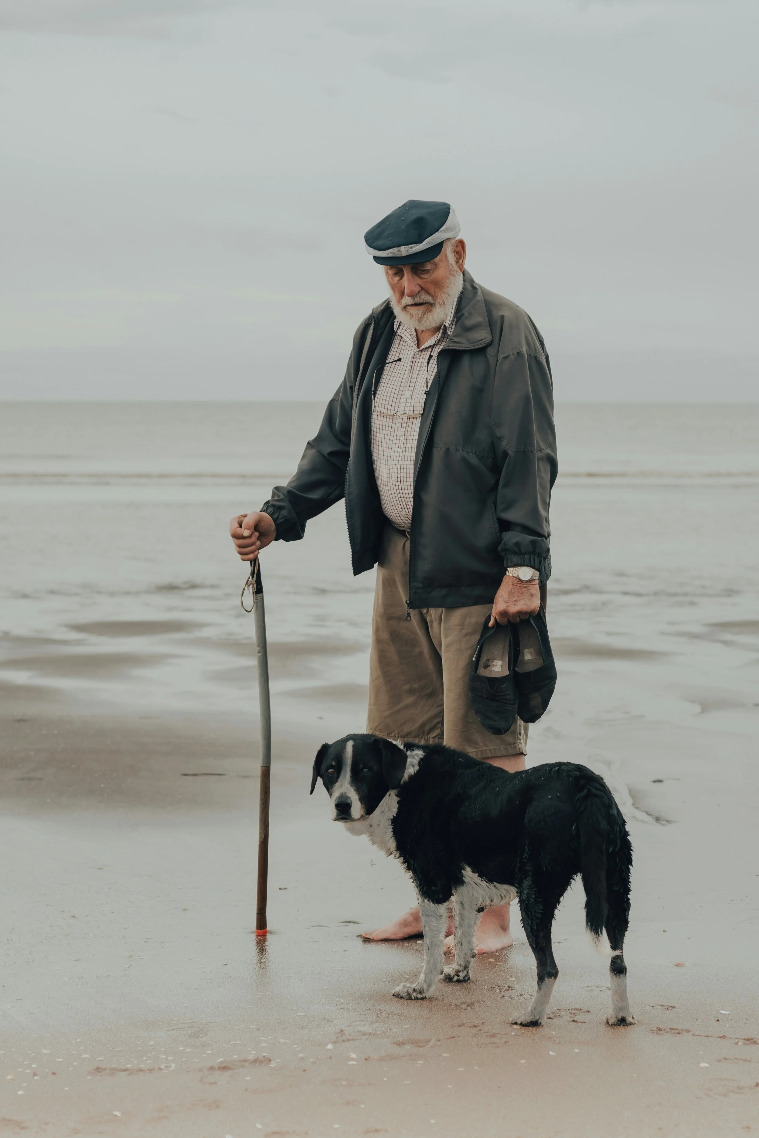 An elderly man standing on a beach with a black and white dog, holding a cane and a cap, wearing a jacket and shorts, under a cloudy sky.