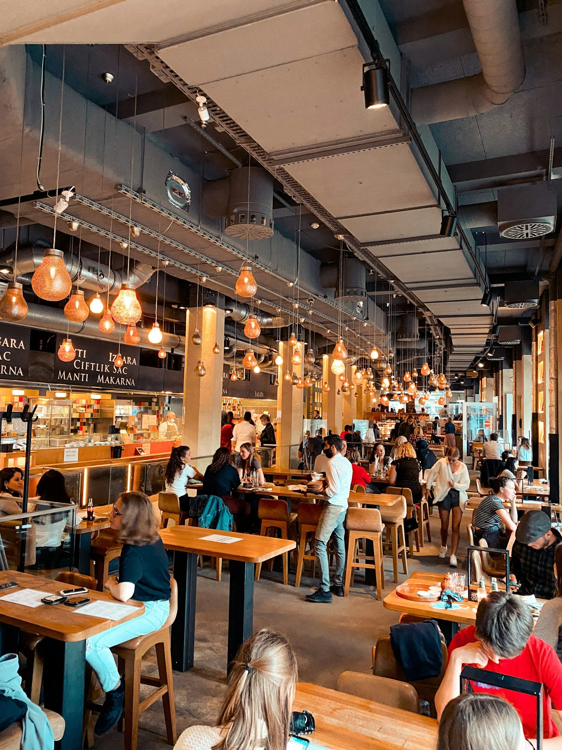 Interior of a busy restaurant with wooden tables and chairs, hanging pendant lights, people dining and waiting, and a visible kitchen area in the background.
