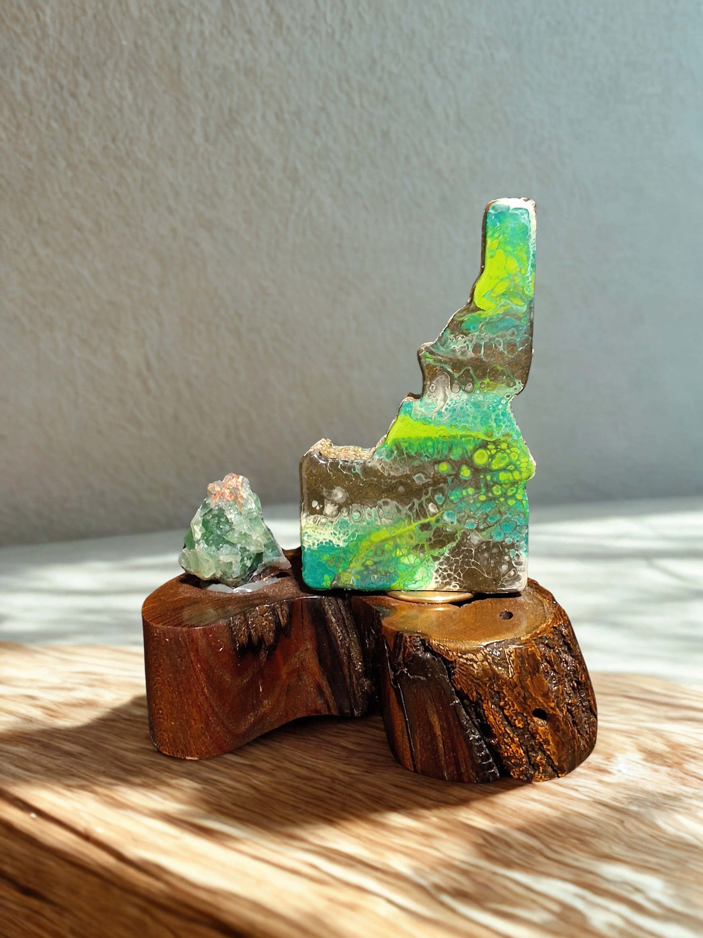 Colorful abstract glass sculpture with green, blue, and yellow hues, mounted on a polished wooden base, with a small glass piece beside it, placed on a wooden surface with soft sunlight and shadow.