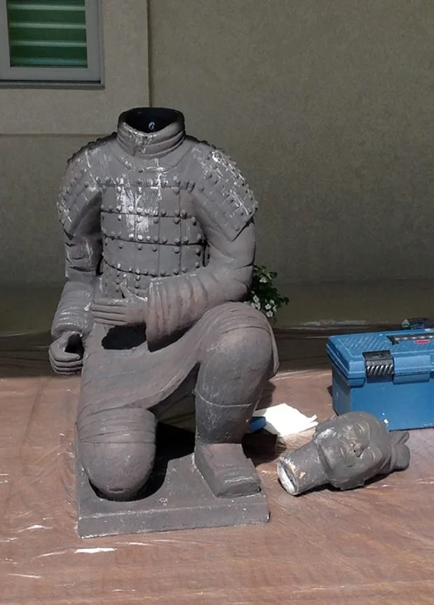 A damaged statue of a kneeling armored figure, missing its head, with the head placed on the floor next to it. The scene is indoors on a wooden floor, with a blue toolbox and some small items nearby.