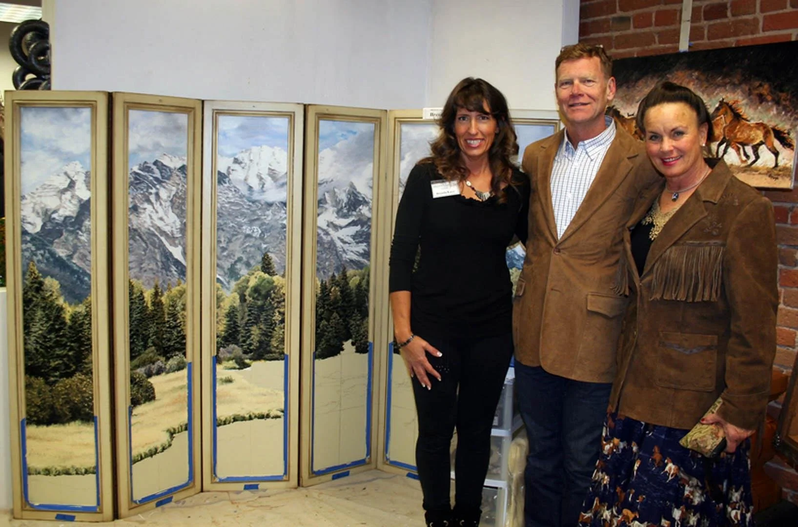 Three people standing together at an indoor art exhibit, with landscape paintings of mountains and trees in the background.