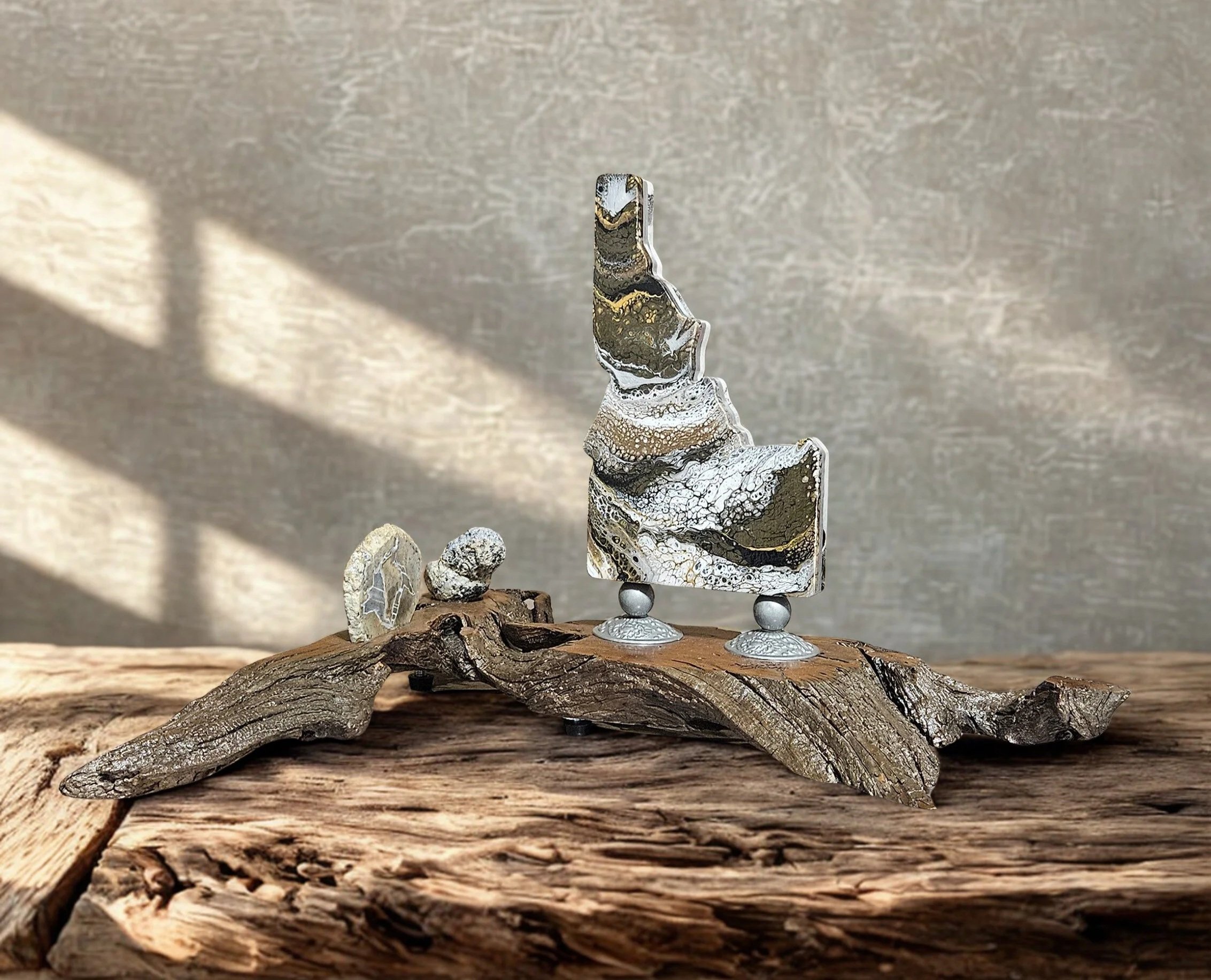 Abstract art sculpture with a multicolored textured piece standing on two small spheres, on a rustic piece of wood, with smaller stones nearby, in a well-lit setting with shadows on a plain background.