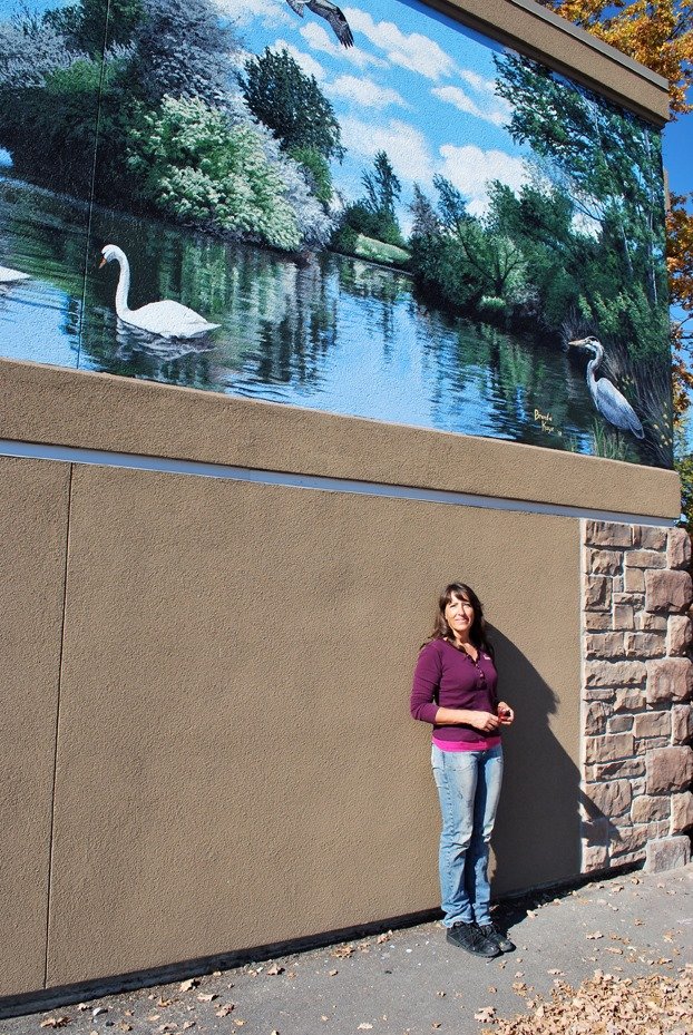 Woman standing outdoors near a building with a large mural of a river scene, including a swan and a heron, with trees and sky in the background.