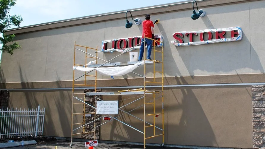 Worker installing a red and black sign on the exterior of a commercial store building, using scaffolding