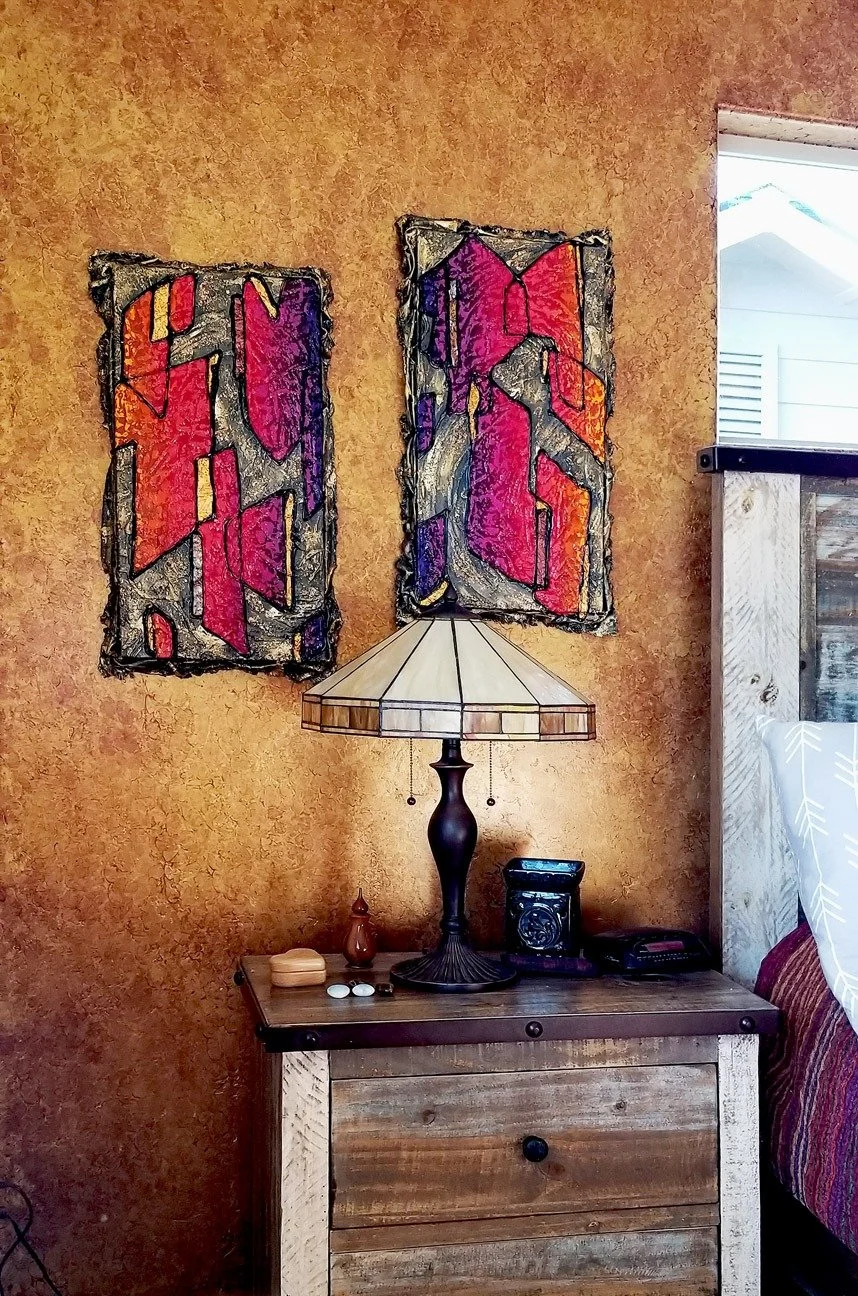 A rustic wooden bedside table with a stained glass lamp on top, decorated with small objects, against an orange textured wall with two stained glass art pieces