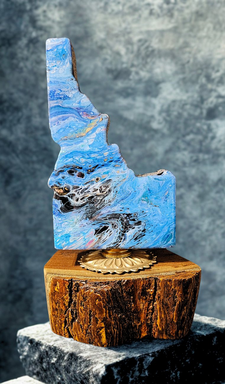 Abstract art sculpture made of blue and black swirling resin or paint, mounted on a wooden base with a brass decorative element, displayed on a stone surface against a textured gray background.