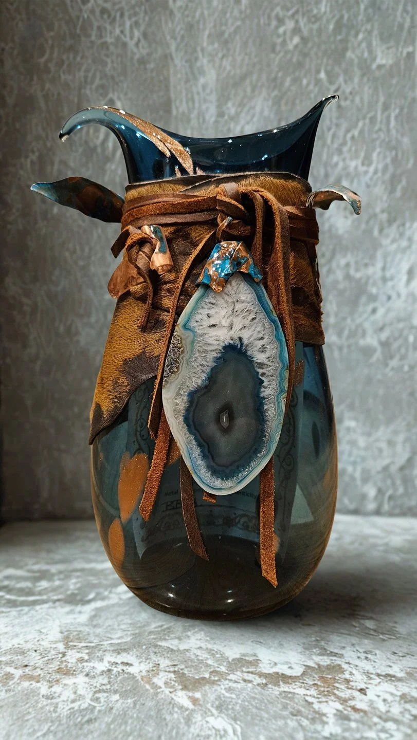 Decorative glass vase with leather and fabric accents, featuring a central agate slice and a dark blue glass top, positioned on a textured surface.