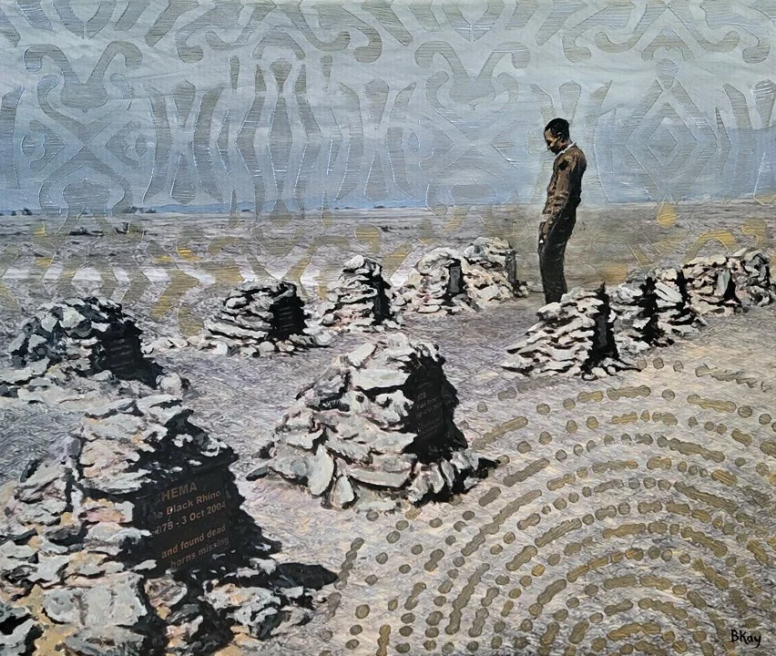 A man stands among heaps of rocks arranged in rows in a desert, with a large patterned mural on the wall behind him.