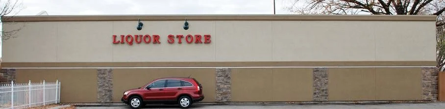 Exterior of a liquor store with a beige wall, stone accents, and a red sign reading 'LIQUOR STORE'; a red SUV parked in front.