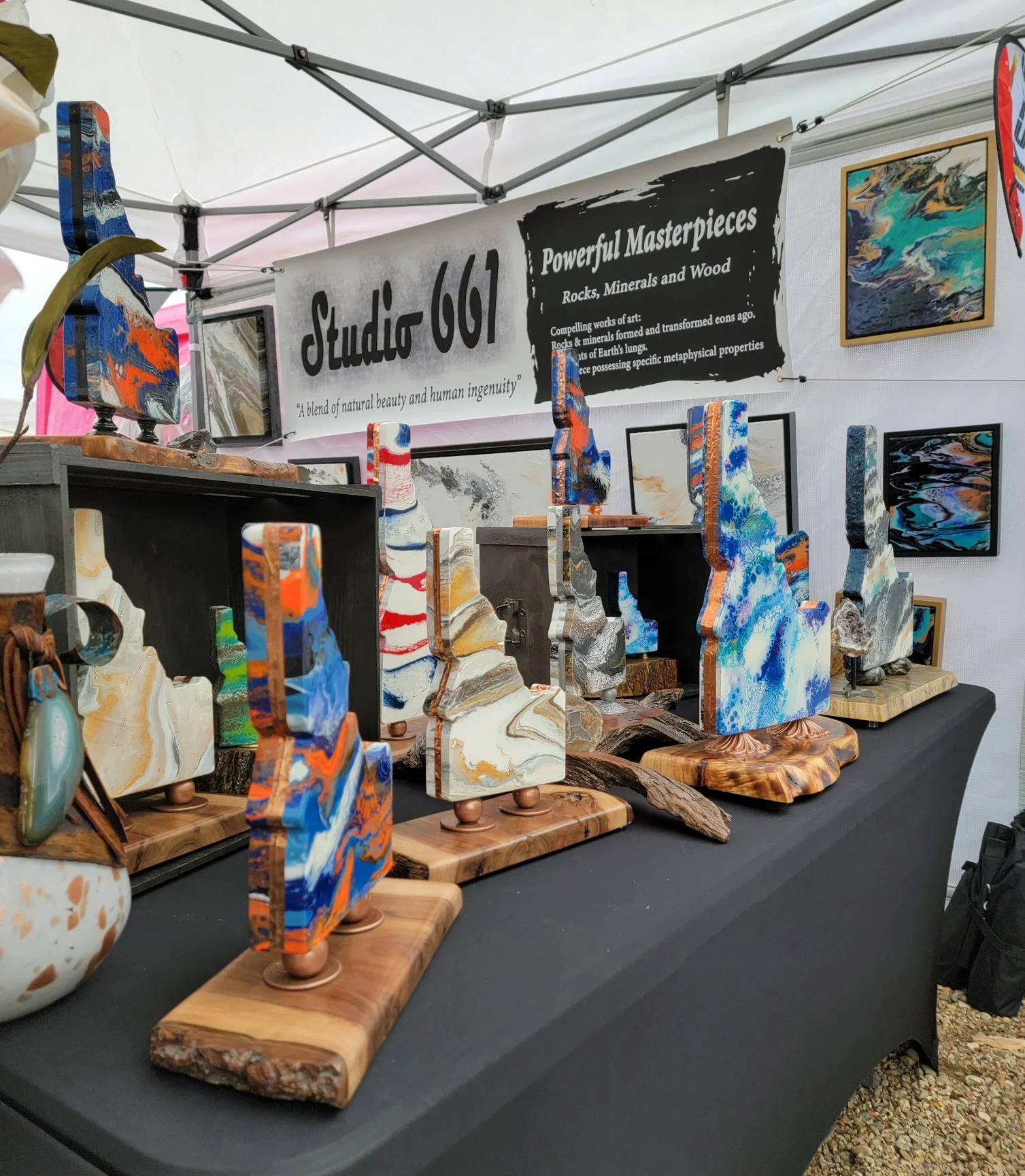 Colorful abstract art pieces on display at a booth with a sign that reads 'Studio 667 Powerfull Masterpieces Rocks, Minerals and Wood.' The art pieces are mounted on wooden bases and arranged on a black table, with framed paintings hanging in the bac